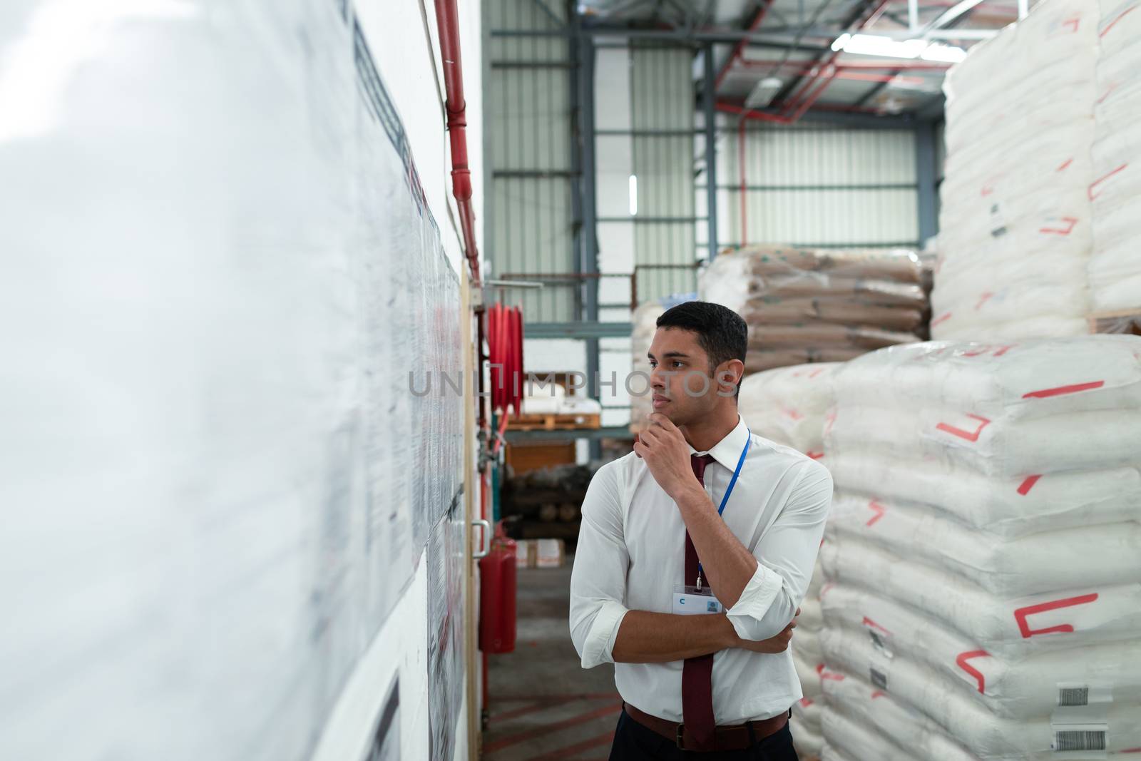 Front view of handsome young mixed-race male supervisor looking at inventory records on wall. This is a freight transportation and distribution warehouse. Industrial and industrial workers concept