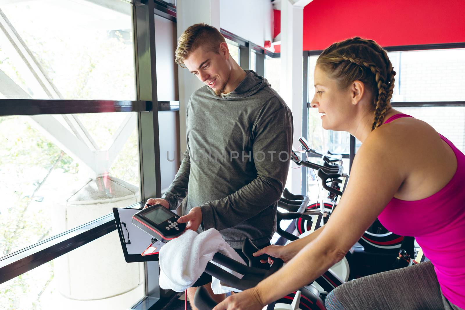 Front view of young Caucasian Male trainer assisting young Caucasian female athlete with exercise in fitness studio. Bright modern gym with fit healthy people working out and training