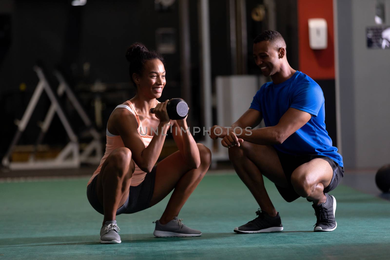 Front view of an African-American woman lifting ball weights while doing squats and an African-American man assisting her  inside a room at a sports centre. Bright modern gym with fit healthy people working out and training. Bright modern gym with fit healthy people working out and training