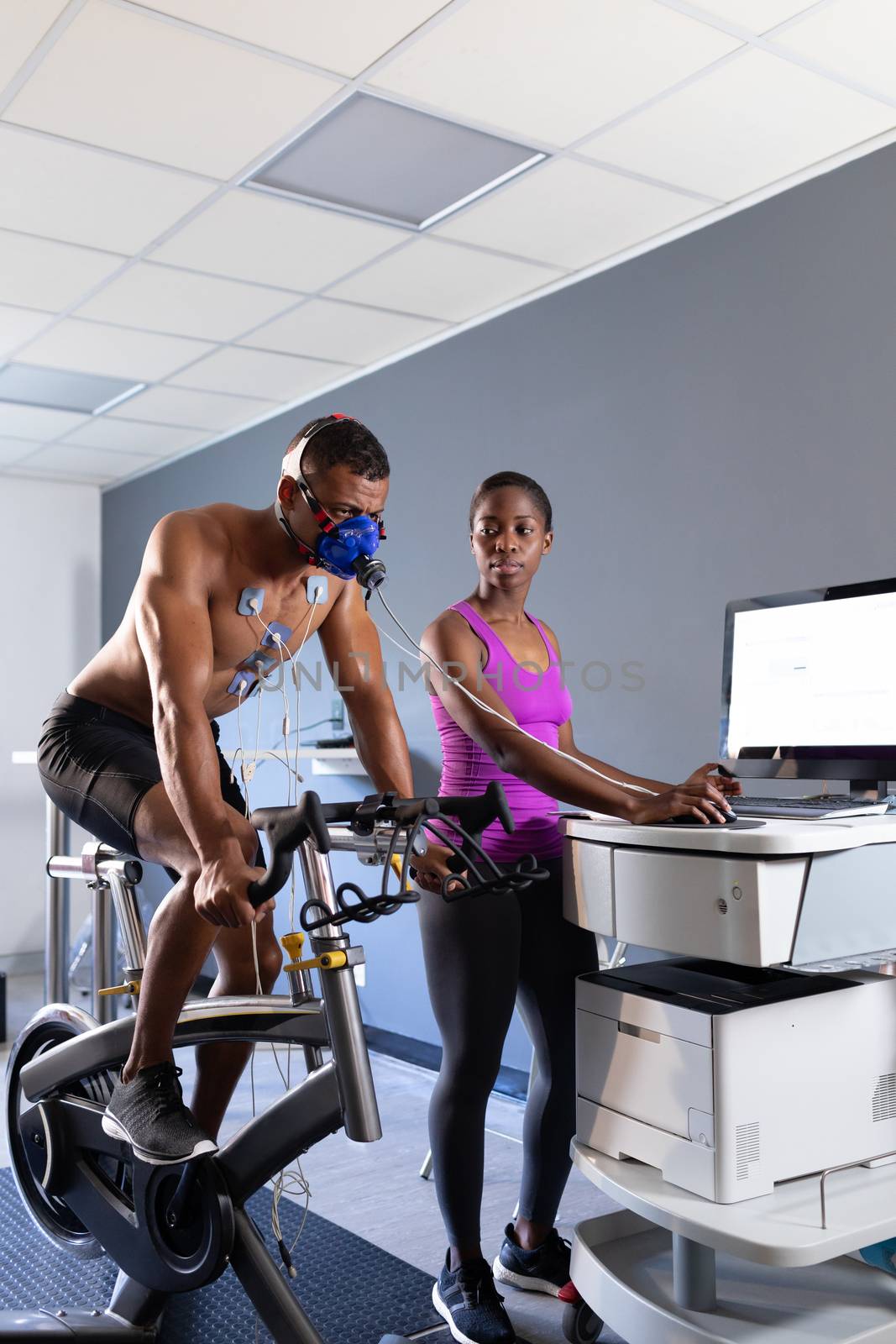 Side view of a naked African-American athletic man doing a fitness test using a mask connected to a monitor while riding an exercise bike and an African-American woman assisting him inside a room at a sports center. Bright modern gym with fit healthy people working out and training. Athlete testing themselves with cardiovascular fitness test on exercise bike