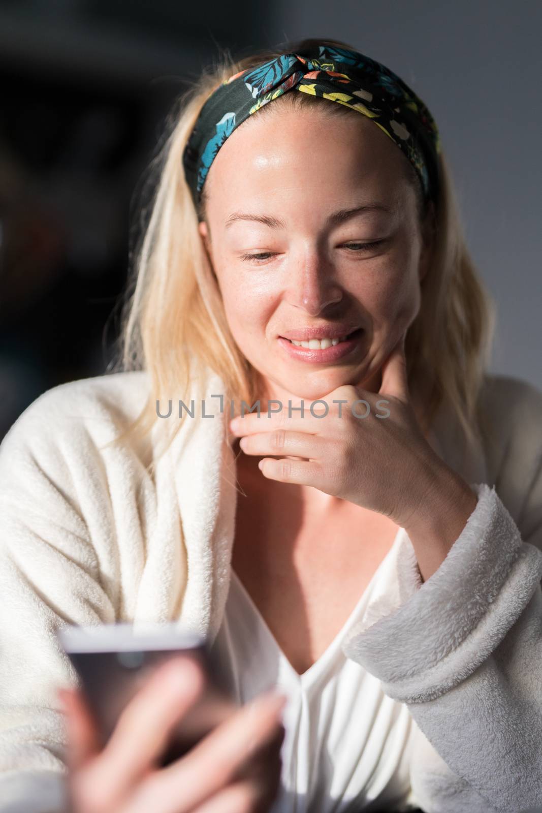 Beautiful caucasian woman at home, feeling comfortable wearing white bathrobe, taking some time to herself, drinking morning coffee and reading news on mobile phone device in the morning.