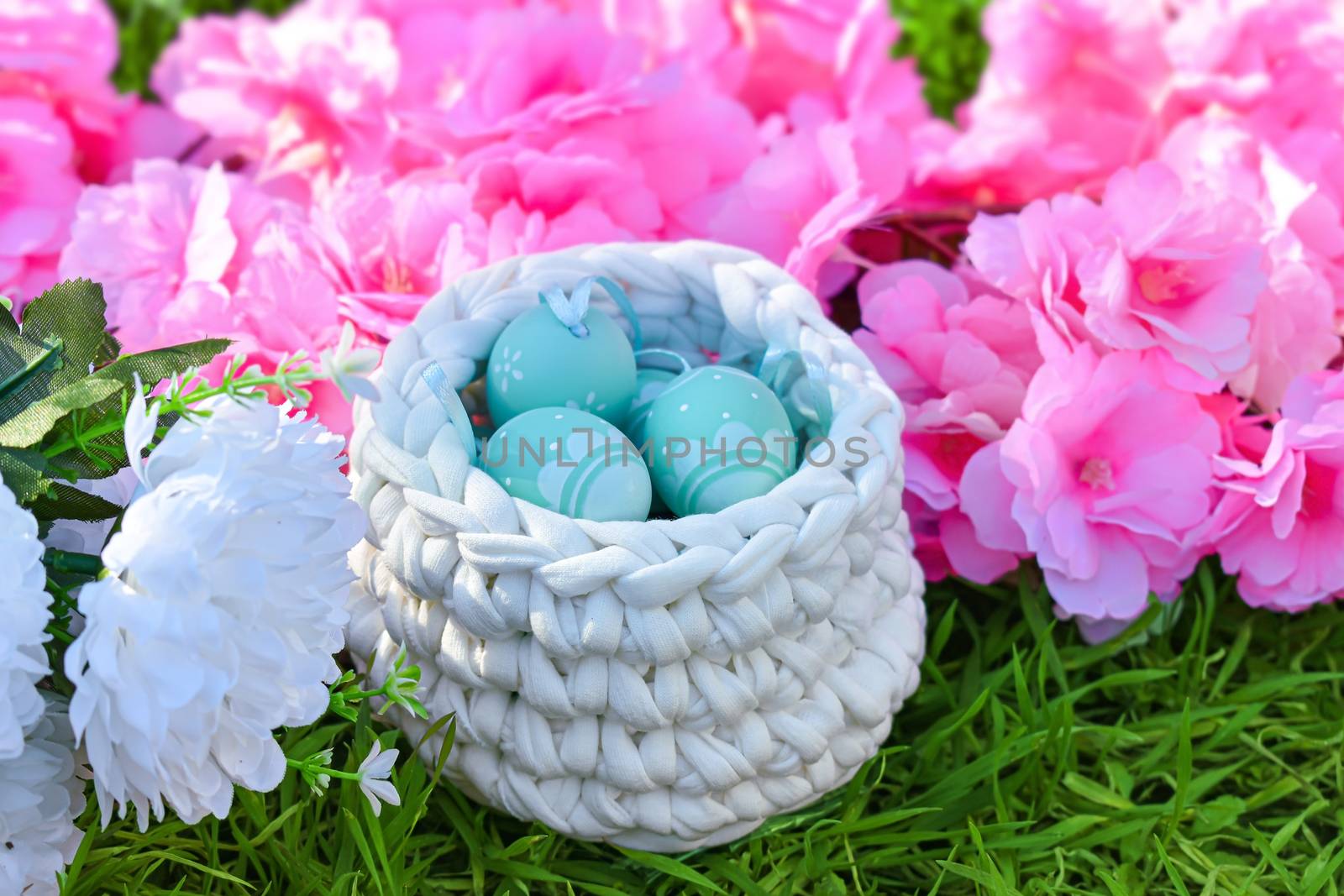 Floral easter background. Painted eggs in crochet basket on green grass. Next to it pink and white flowers decoration.