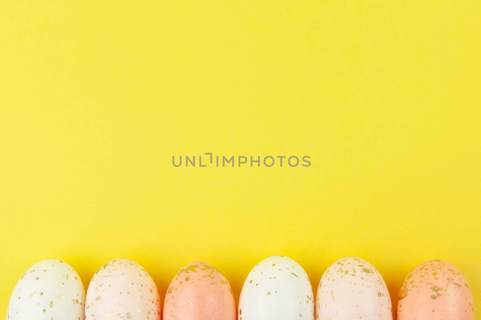 Creatively painted eggs in pastel colors decorated with gold leaf are arranged in row along bottom edge on yellow background, copy space. Happy Easter DIY concept. Flat lay. Horizontal.