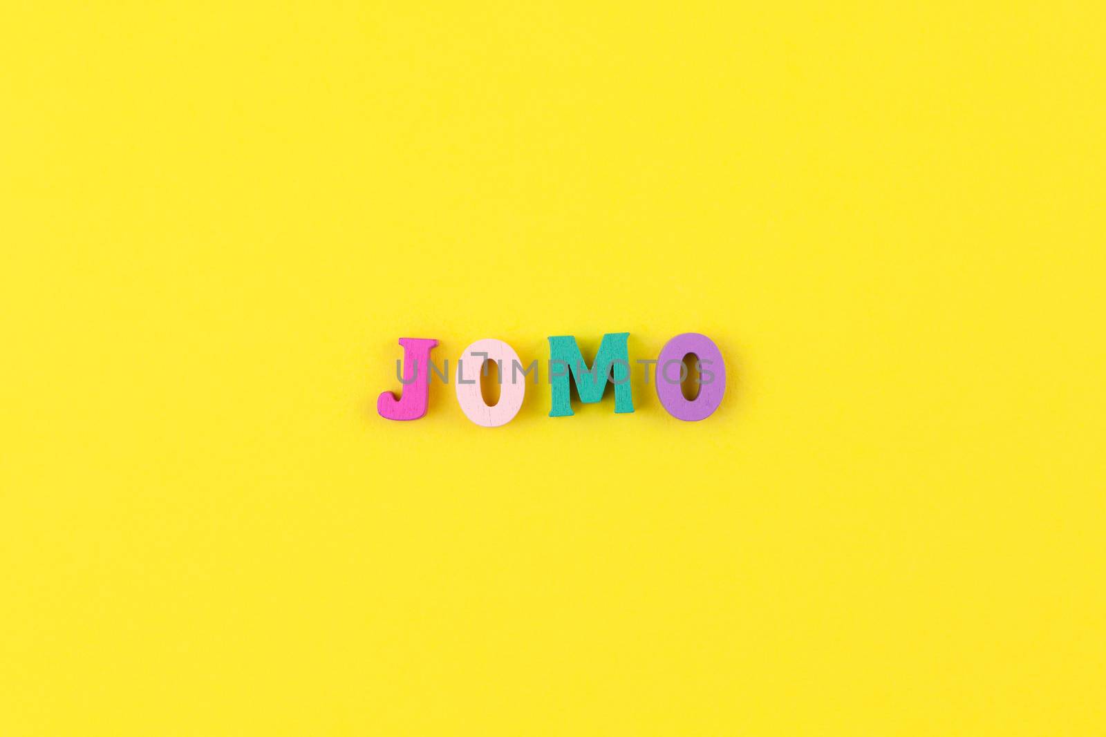 Abbreviation word JOMO in multicolored wooden letters on pastel yellow background. JOMO - Joy Of Missing Out. Opposition, choice, social problem, digital detox. Flat lay, copy space, minimalism style by ALLUNEED