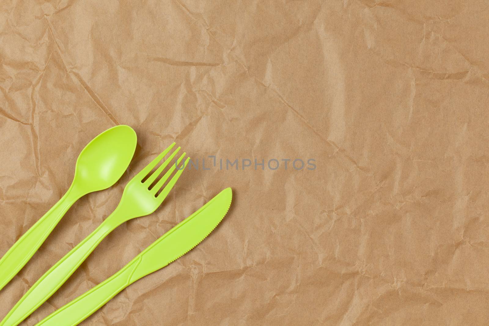 Reusable recyclable green fork, spoon, knife made from corn starch on brown crumpled craft paper, copy space. Eco, zero waste, alternative to plastic concept. Flat lay. Horizontal. Closeup.