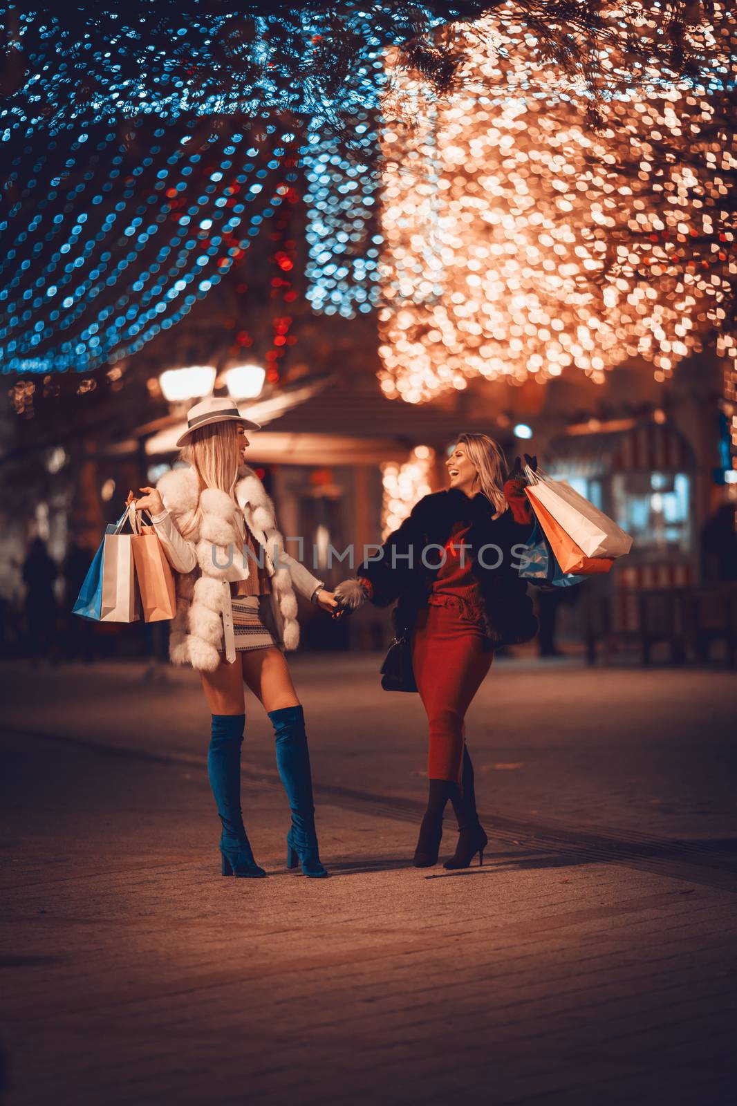 Young beautiful cheerful two sisters with colorful shopping bags having fun and walking in the city street at night at Christmas time.