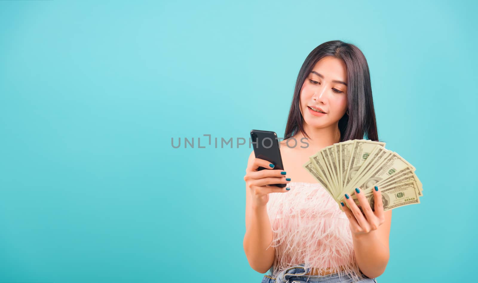 Smiling face portrait asian beautiful woman her usring mobile smart phone and holding bunch money looking at smartphone on blue background, with copy space for text