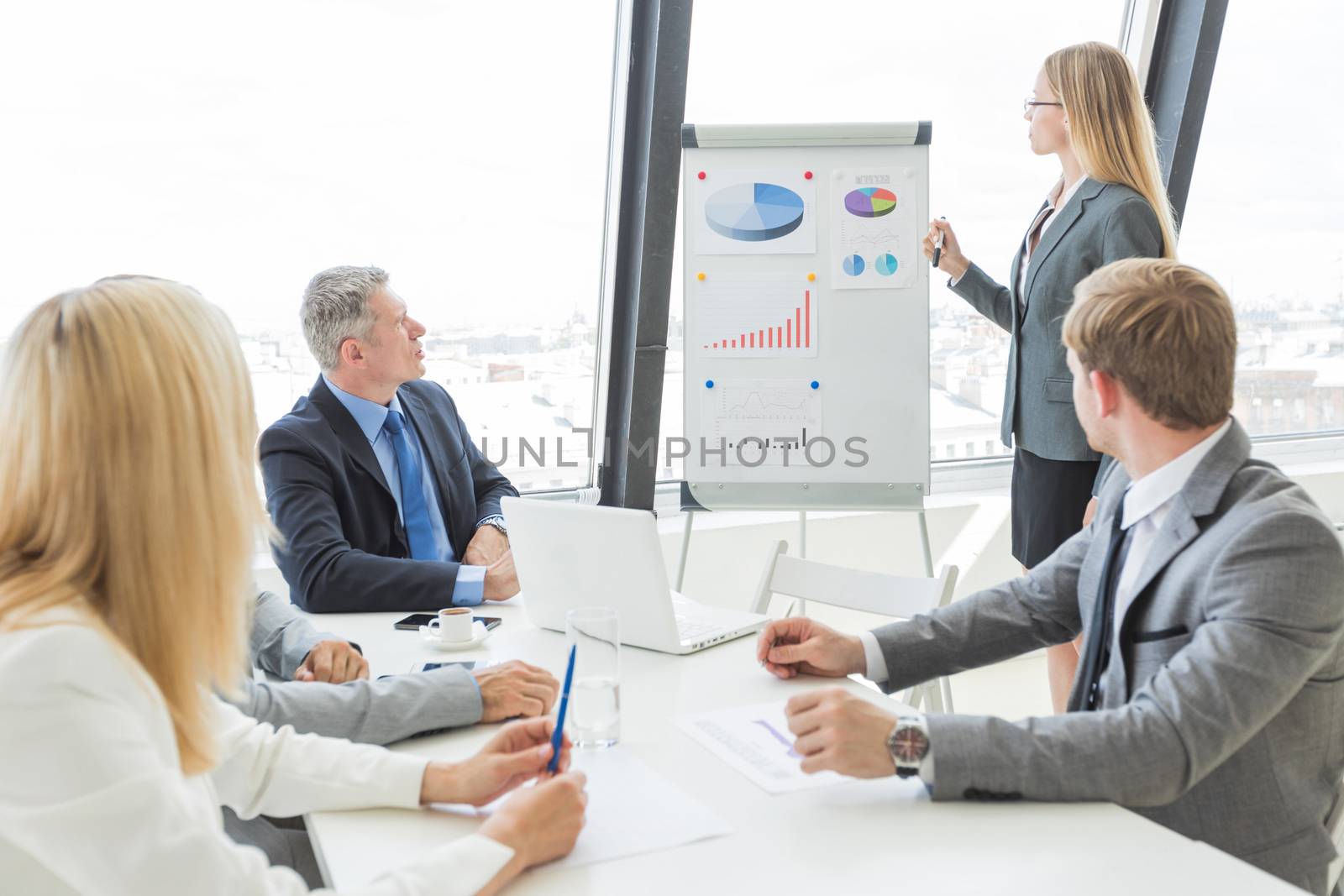 Businesswoman giving presentation using financial diagrams at flipchart to colleagues in office at meeting