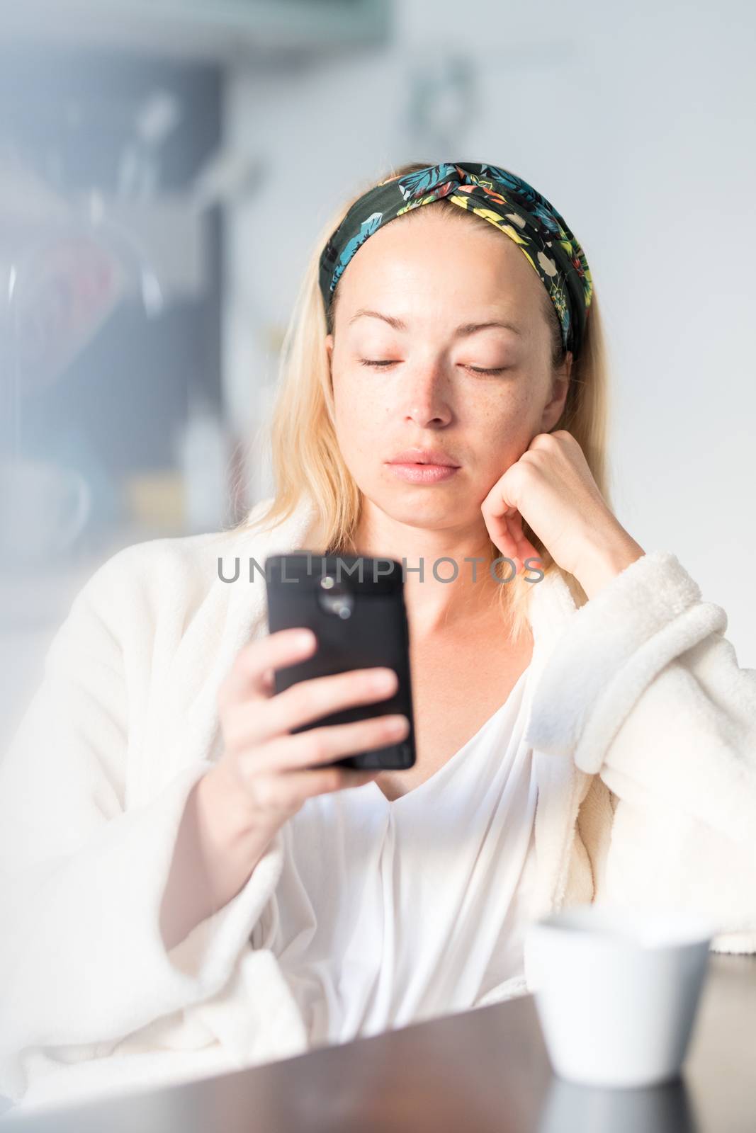 Beautiful caucasian woman at home, feeling comfortable wearing white bathrobe, taking some time to herself, drinking morning coffee and reading news on mobile phone device in the morning.