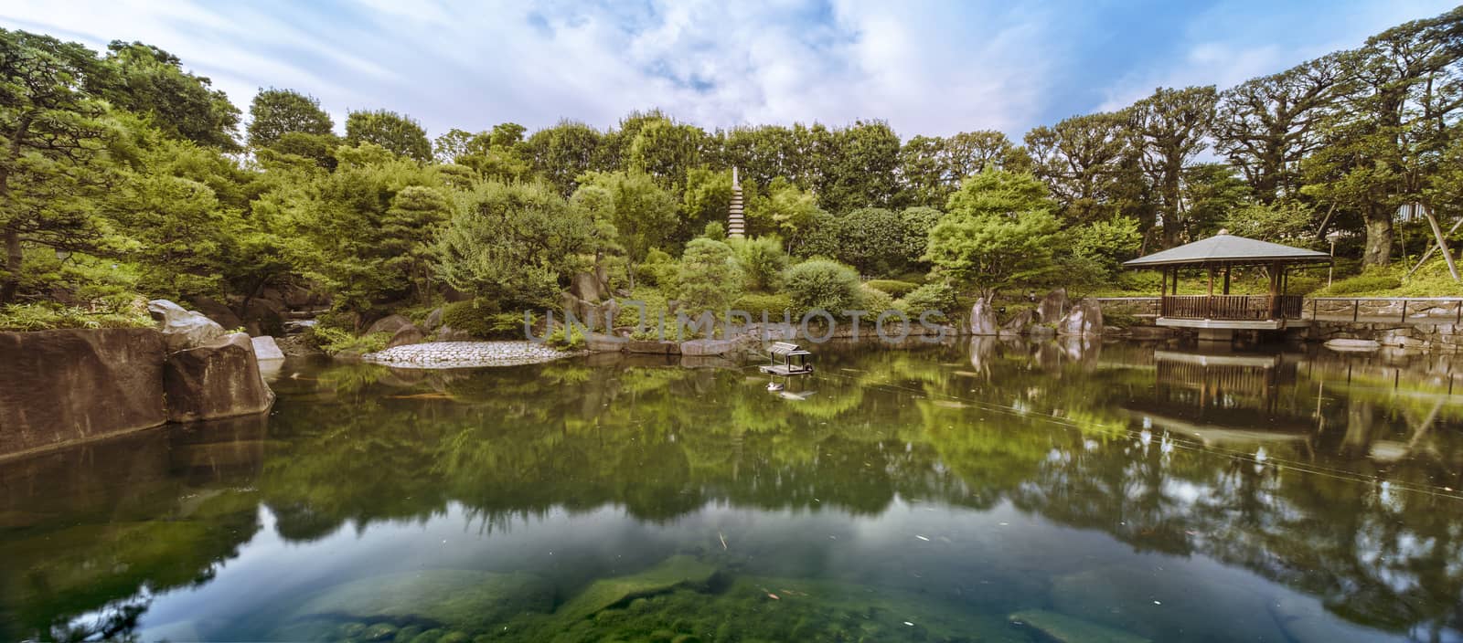 Central pond of Mejiro Garden which is surrounded by large flat stones under the foliage of the momiji maple trees and pines. The sky is reflected symetrically in the water of the pond.