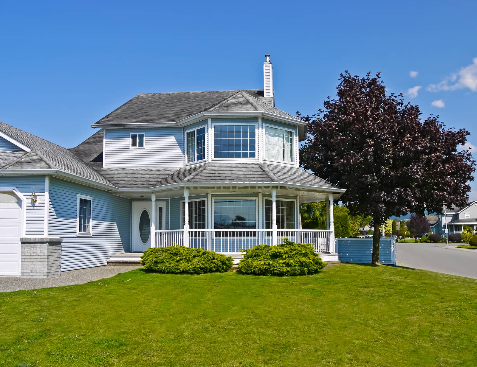 Large residential house with surrounding porch and big green lawn at the entrance. Big family home with black cherry tree beside on blue sky background