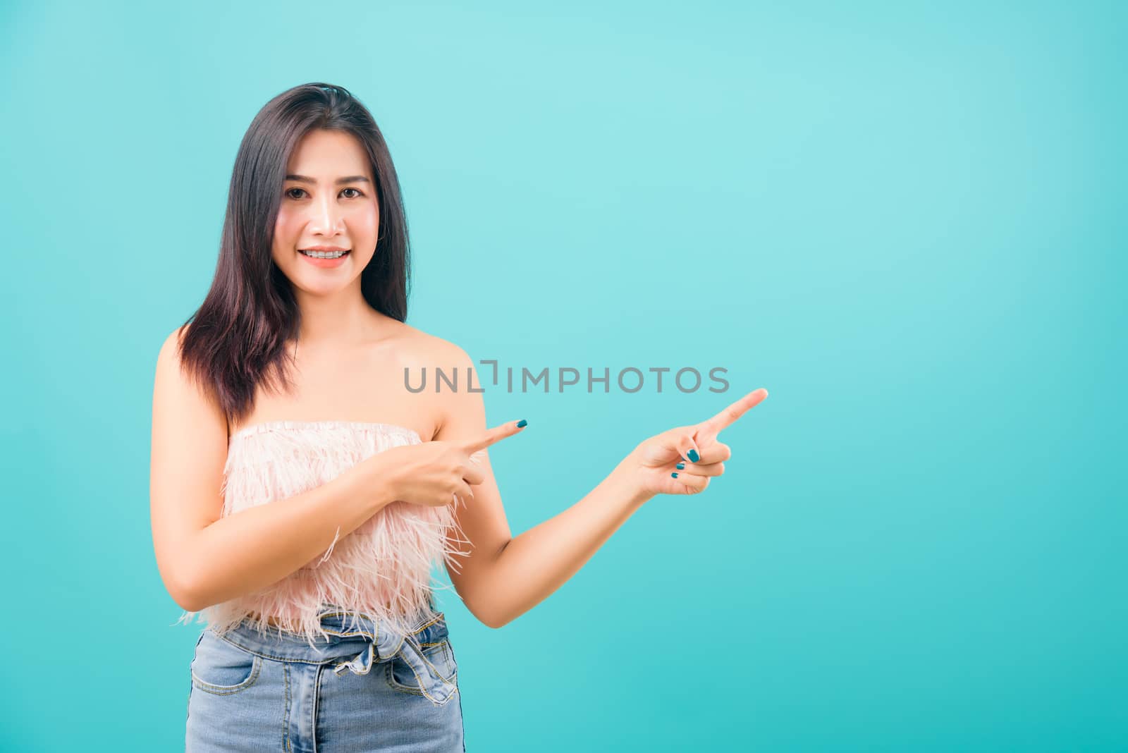 Portrait asian beautiful woman smiling looking camera her standing pointing finger out on blue background, with copy space for text