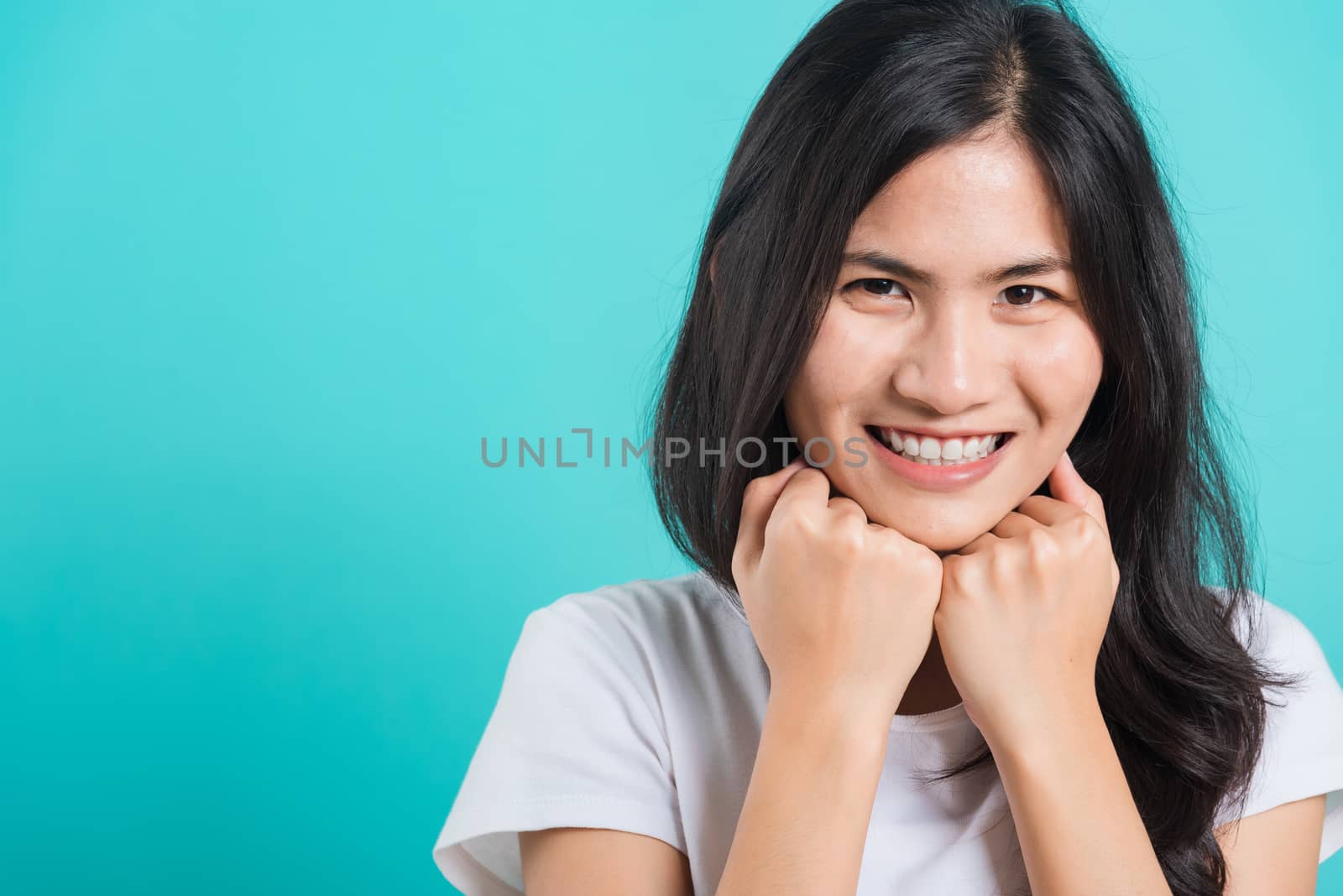 Portrait Asian beautiful happy young woman wear t-shirt glad keeps both hands under chin smiles pleasantly, on blue background, with copy space