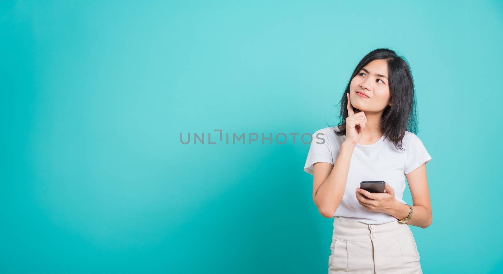 Portrait Asian beautiful happy young woman standing smile, posing using mobile phone her thinking dreaming on blue background, with copyspace