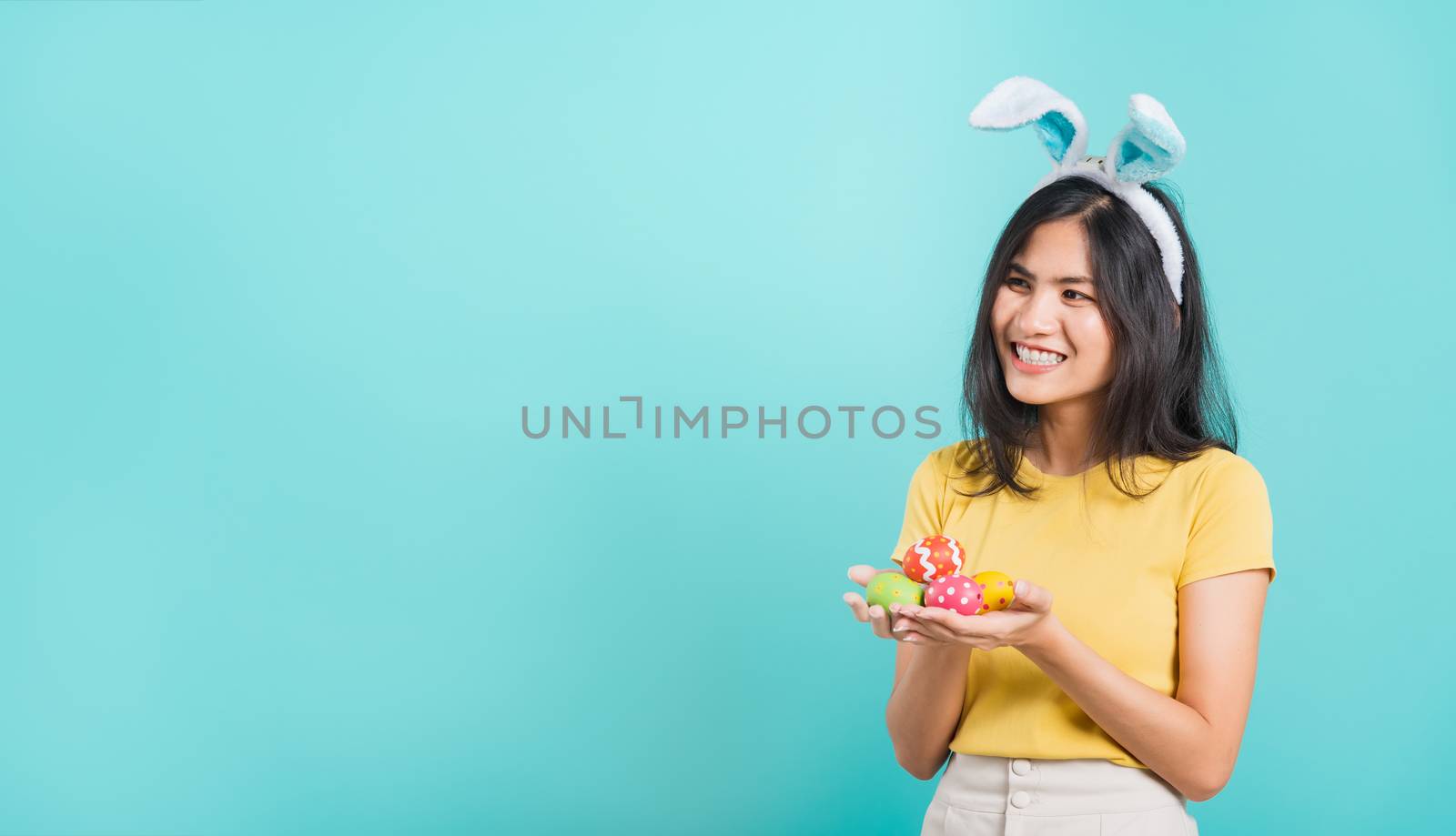 Portrait Asian beautiful happy young woman smile white teeth wear yellow t-shirt standing with bunny ears and holding Easter eggs looking to space, on a blue background with copy space