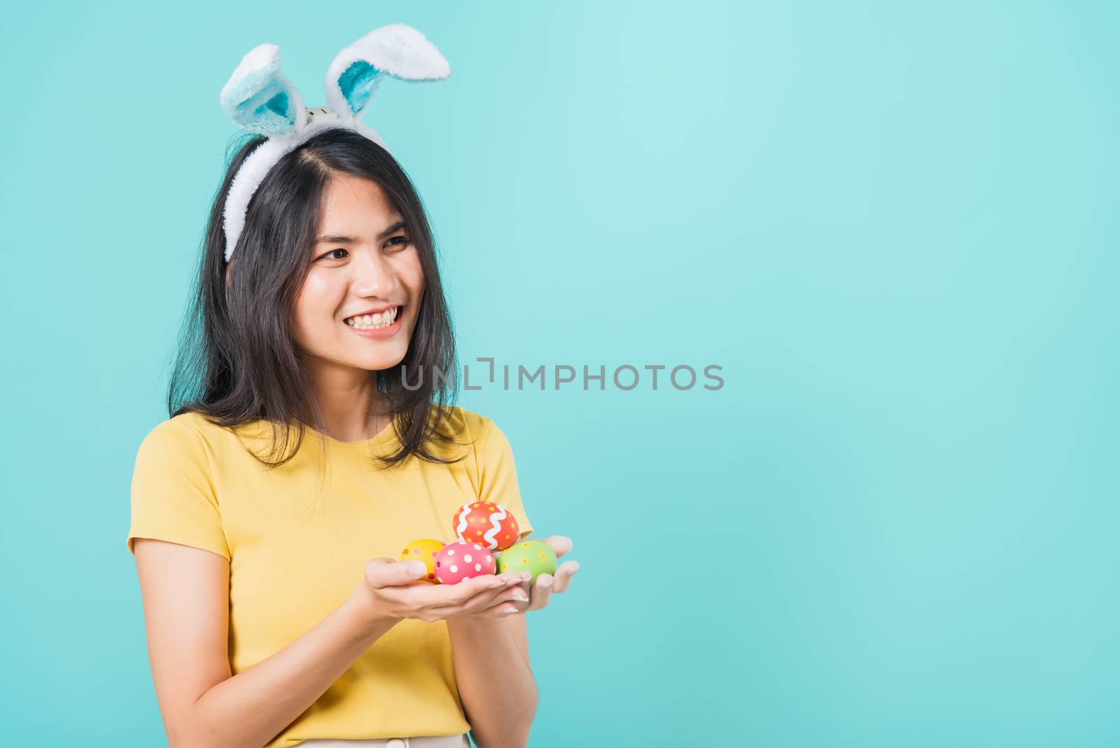 Portrait Asian beautiful happy young woman smile white teeth wear yellow t-shirt standing with bunny ears and holding Easter eggs looking to space, on a blue background with copy space