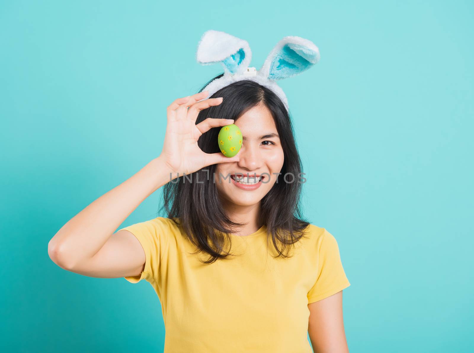 Portrait Asian beautiful happy young woman smile white teeth wear yellow t-shirt standing with bunny ears and holding Easter eggs near the eye, on blue background with copy space