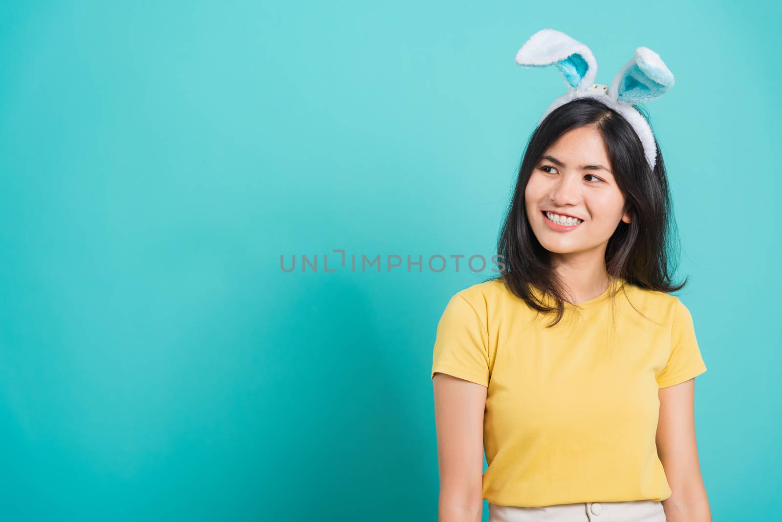 Portrait Asian beautiful happy young woman smile white teeth wear yellow t-shirt standing with bunny ears her looking to space, on blue background with copy space