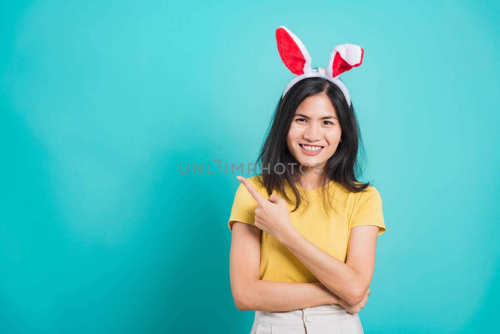 Portrait Asian beautiful happy young woman smile white teeth wear yellow t-shirt standing with bunny ears pointing to space her looking camera, on blue background with copy space