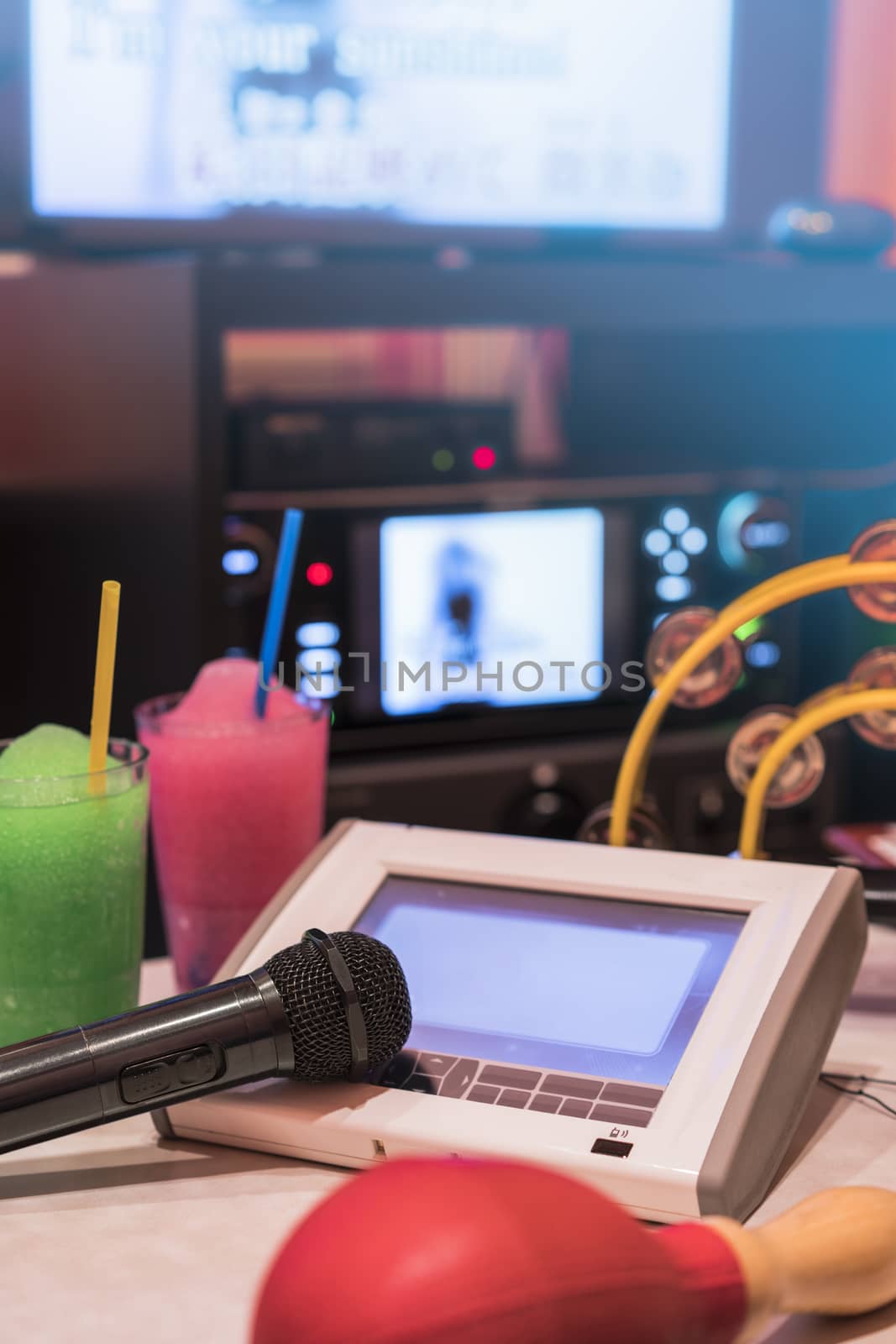 Black microphone in karaoke club, with remote controller, melon and strawberry soda drinks, yellow tambourine and screen for singing music on stage party. 