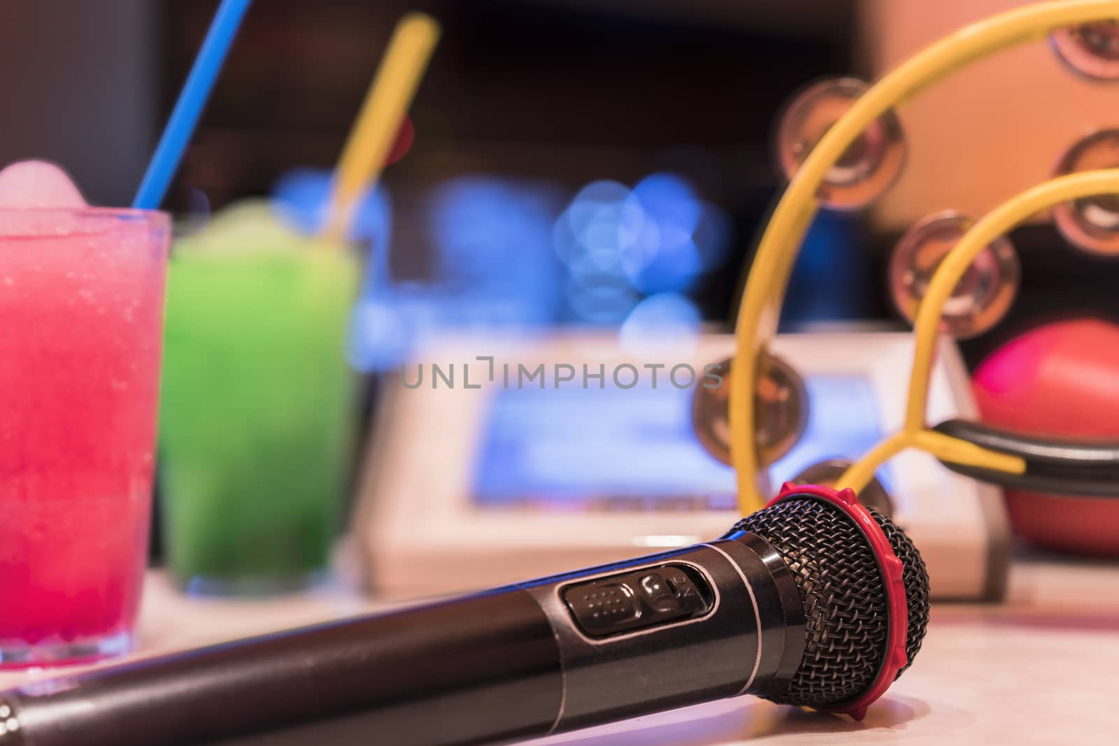 Black microphone in karaoke club, with remote controller, melon and strawberry soda drinks, yellow tambourine and screen for singing music on stage party. 