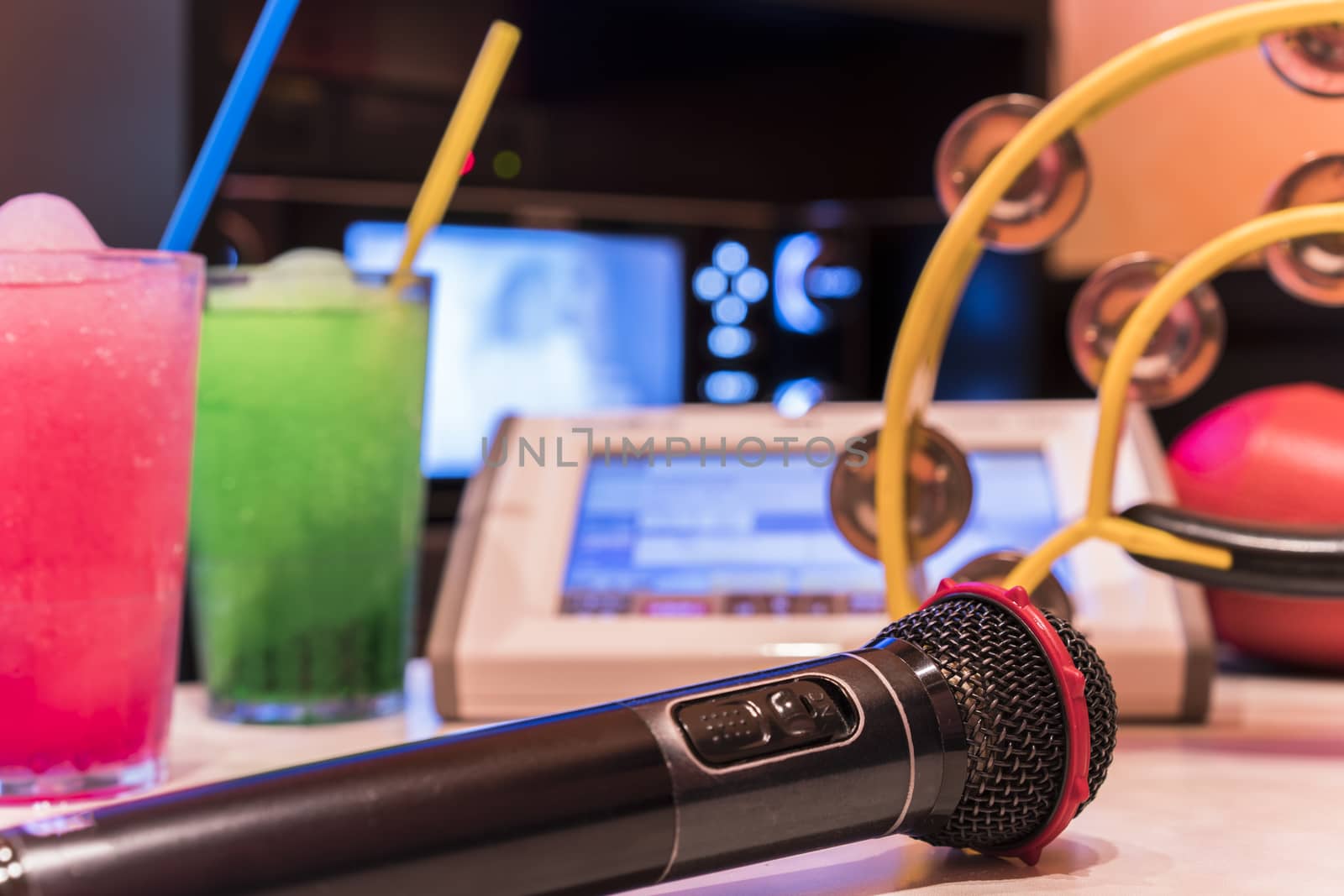 Black microphone in karaoke club, with remote controller, melon and strawberry soda drinks, yellow tambourine and screen for singing music on stage party. 