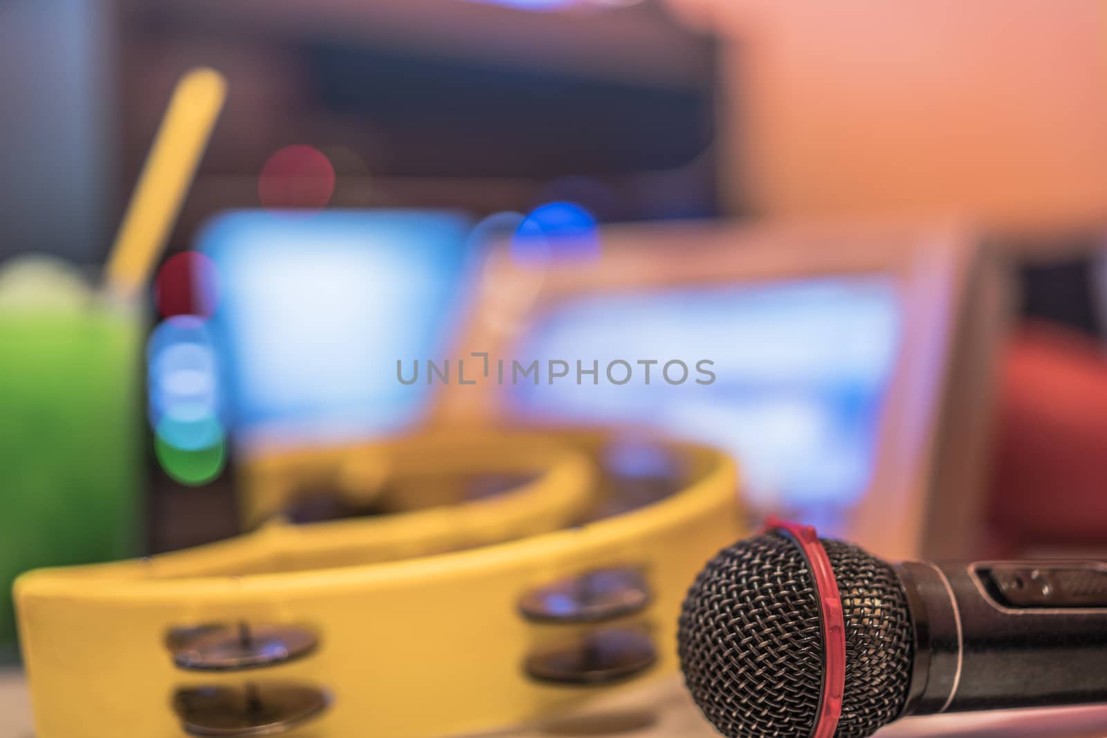 Black microphone in karaoke club, with remote controller, melon and strawberry soda drinks, yellow tambourine and screen for singing music on stage party. 