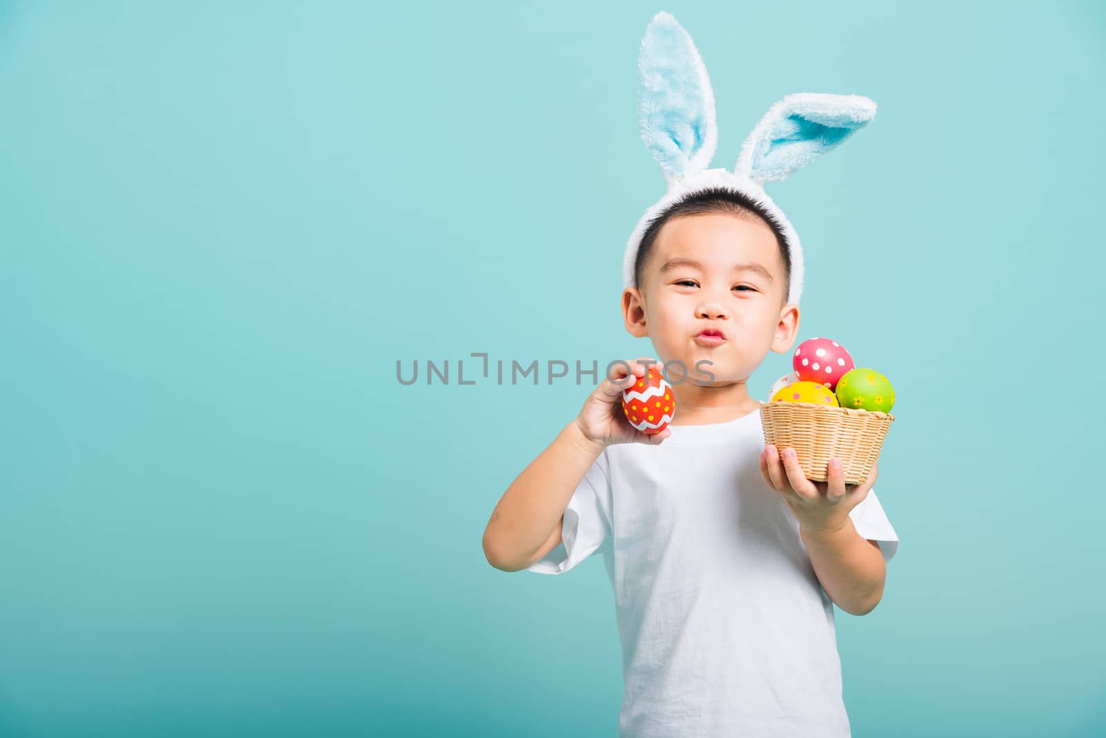 Asian cute little child boy smile beaming wearing bunny ears and a white T-shirt, standing to hold a basket with full Easter eggs. And other hand holds an easter egg on blue background with copy space