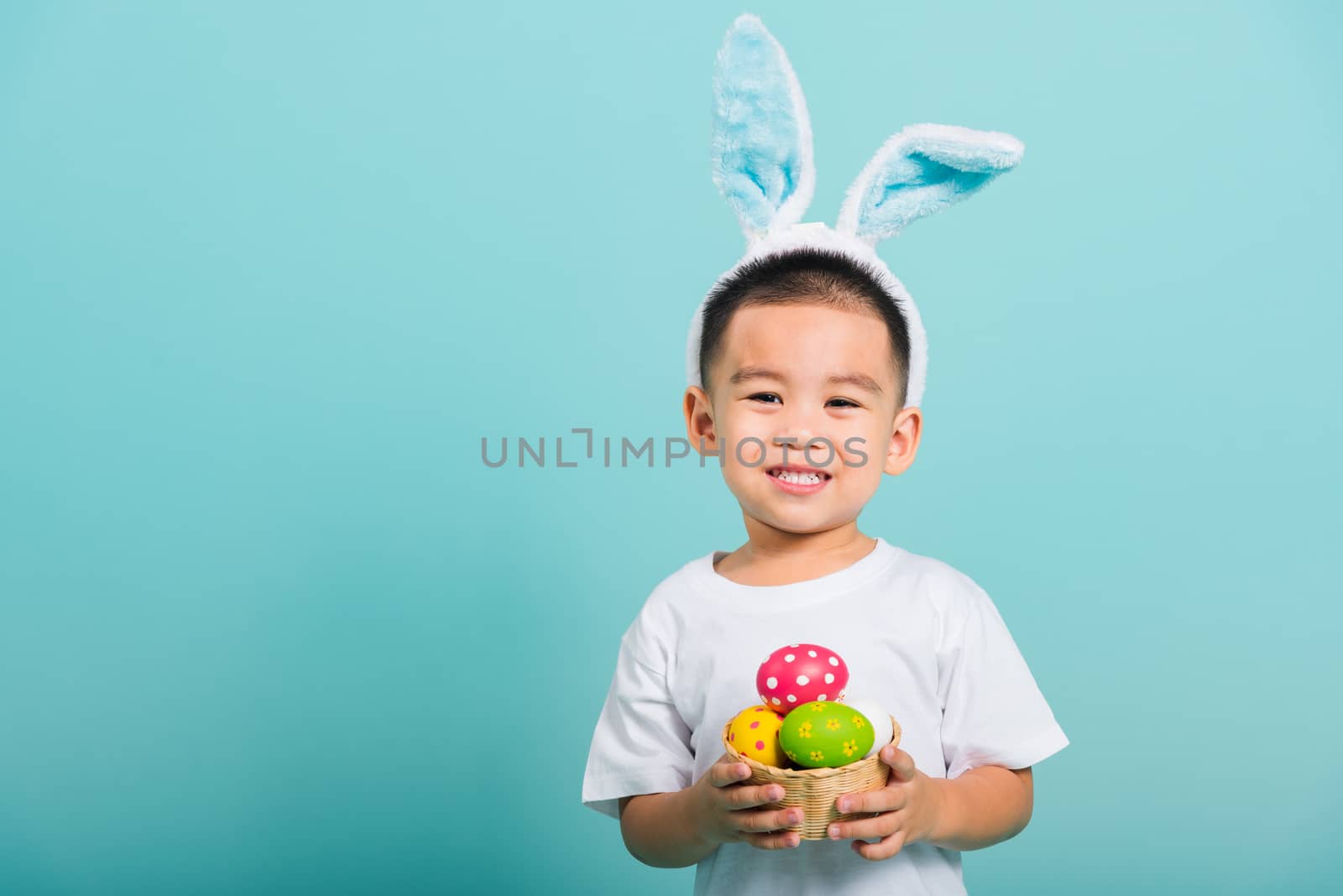 Asian cute little child boy smile beaming wearing bunny ears and a white T-shirt, standing to hold a basket with full Easter eggs on blue background with copy space