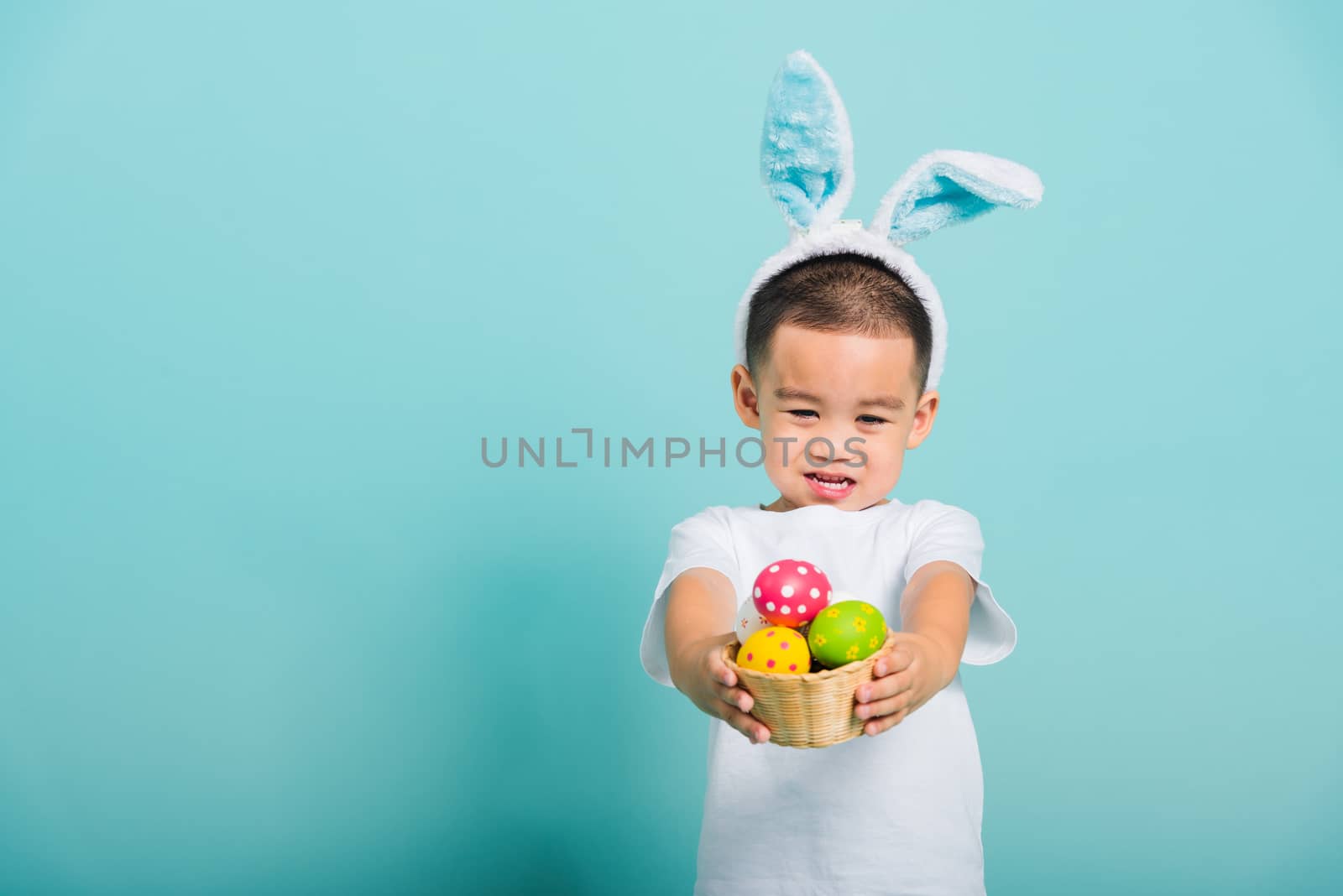 Asian cute little child boy smile beaming wearing bunny ears and a white T-shirt, standing to hold a basket with full Easter eggs on blue background with copy space