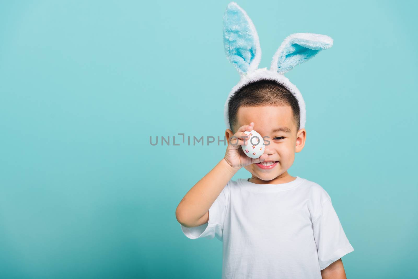 Asian cute little child boy smile beaming wearing bunny ears and a white T-shirt, standing to holds colored easter eggs instead of eyes on blue background with copy space for text