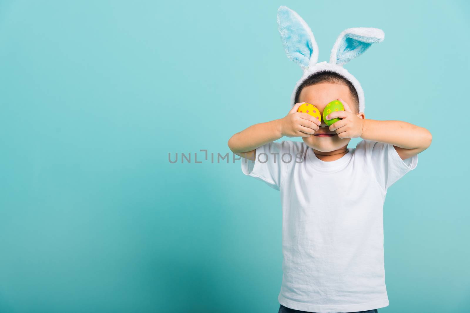 Asian cute little child boy smile beaming wearing bunny ears and a white T-shirt, standing to holds colored easter eggs instead of eyes on blue background with copy space for text