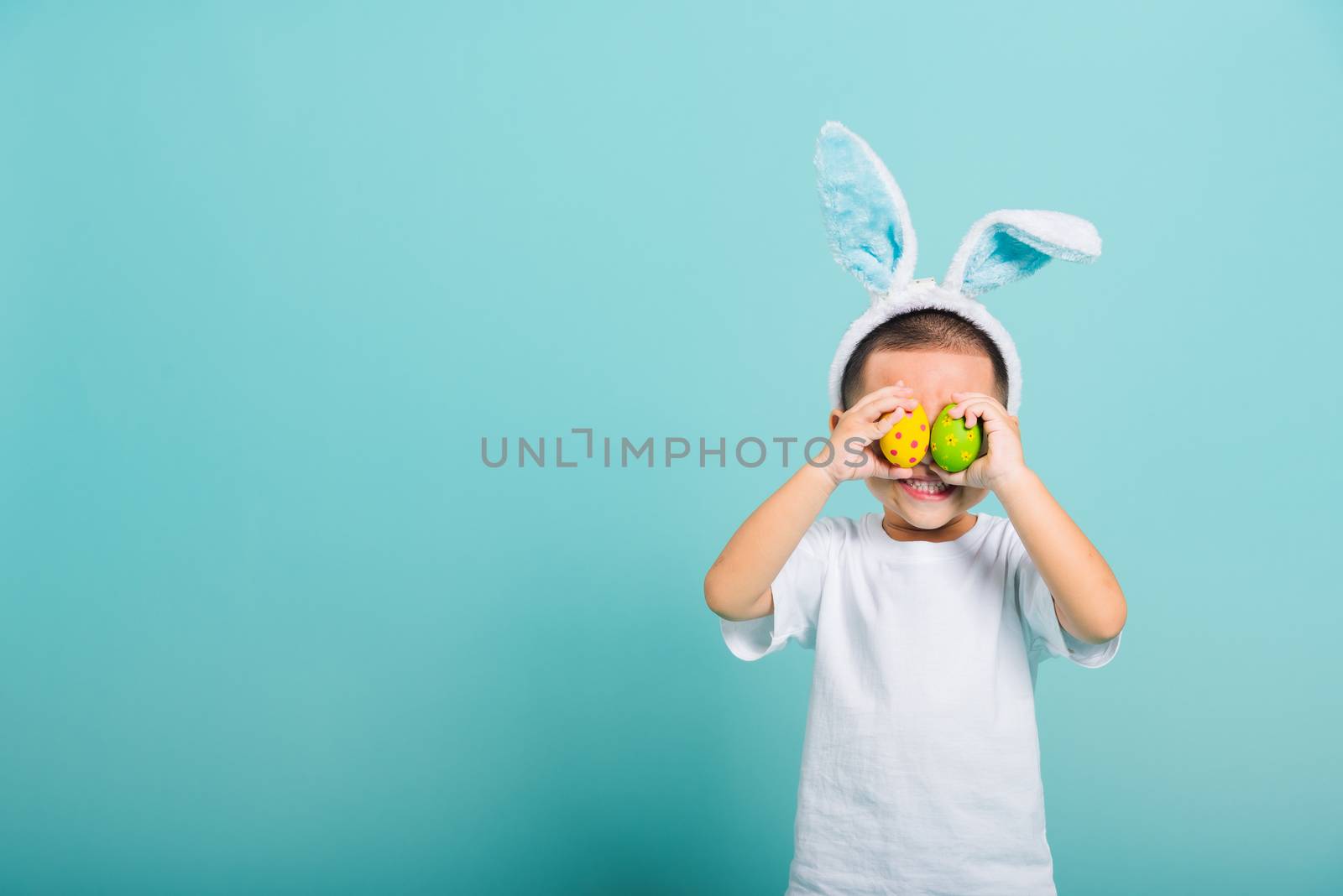 Asian cute little child boy smile beaming wearing bunny ears and a white T-shirt, standing to holds colored easter eggs instead of eyes on blue background with copy space for text