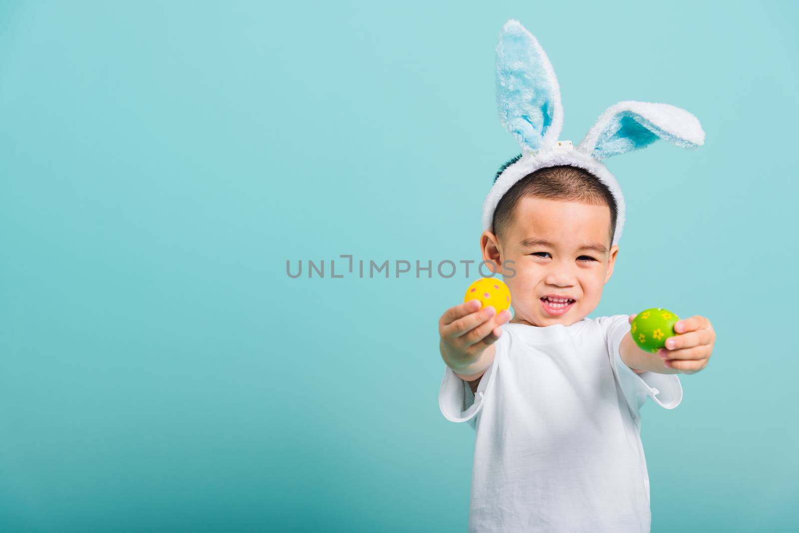 Asian cute little child boy smile beaming wearing bunny ears and a white T-shirt, standing using hand holding and show colored easter eggs on blue background with copy space