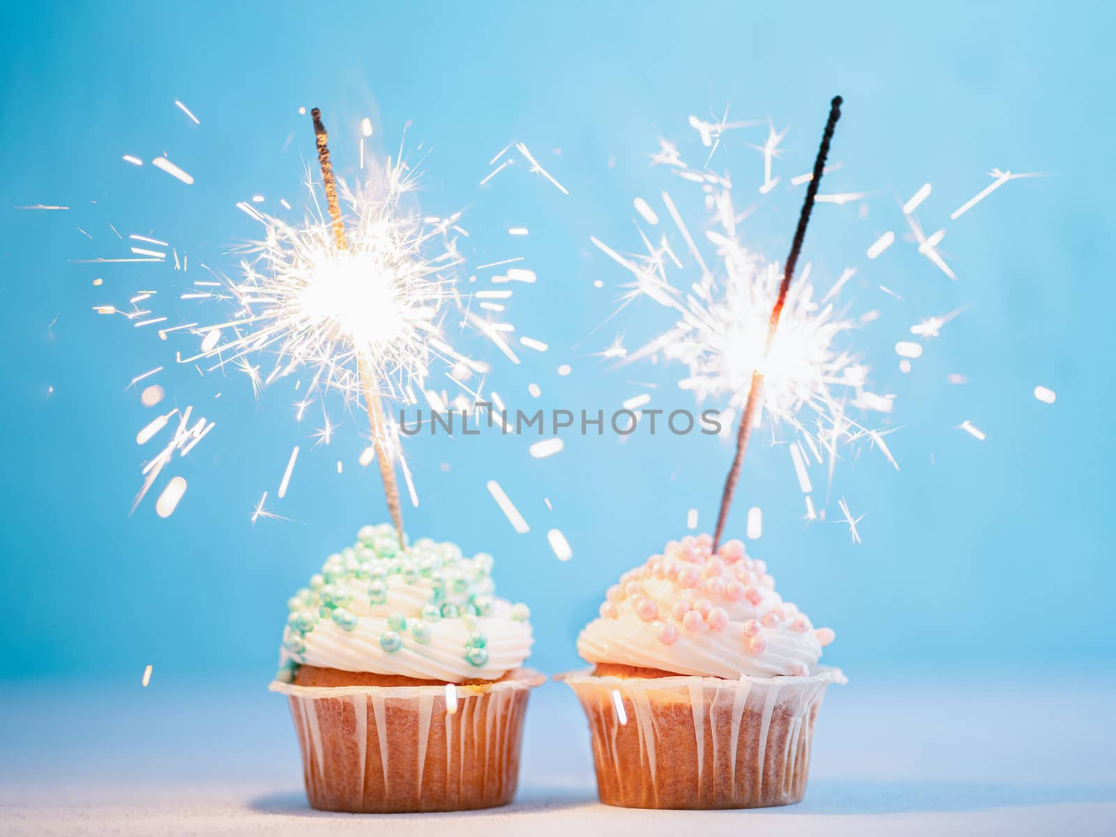 Two cupcakes decorated with colorful sprinkles and sparklers. Festive cupcakes with blue and pink sprinkles on blue background