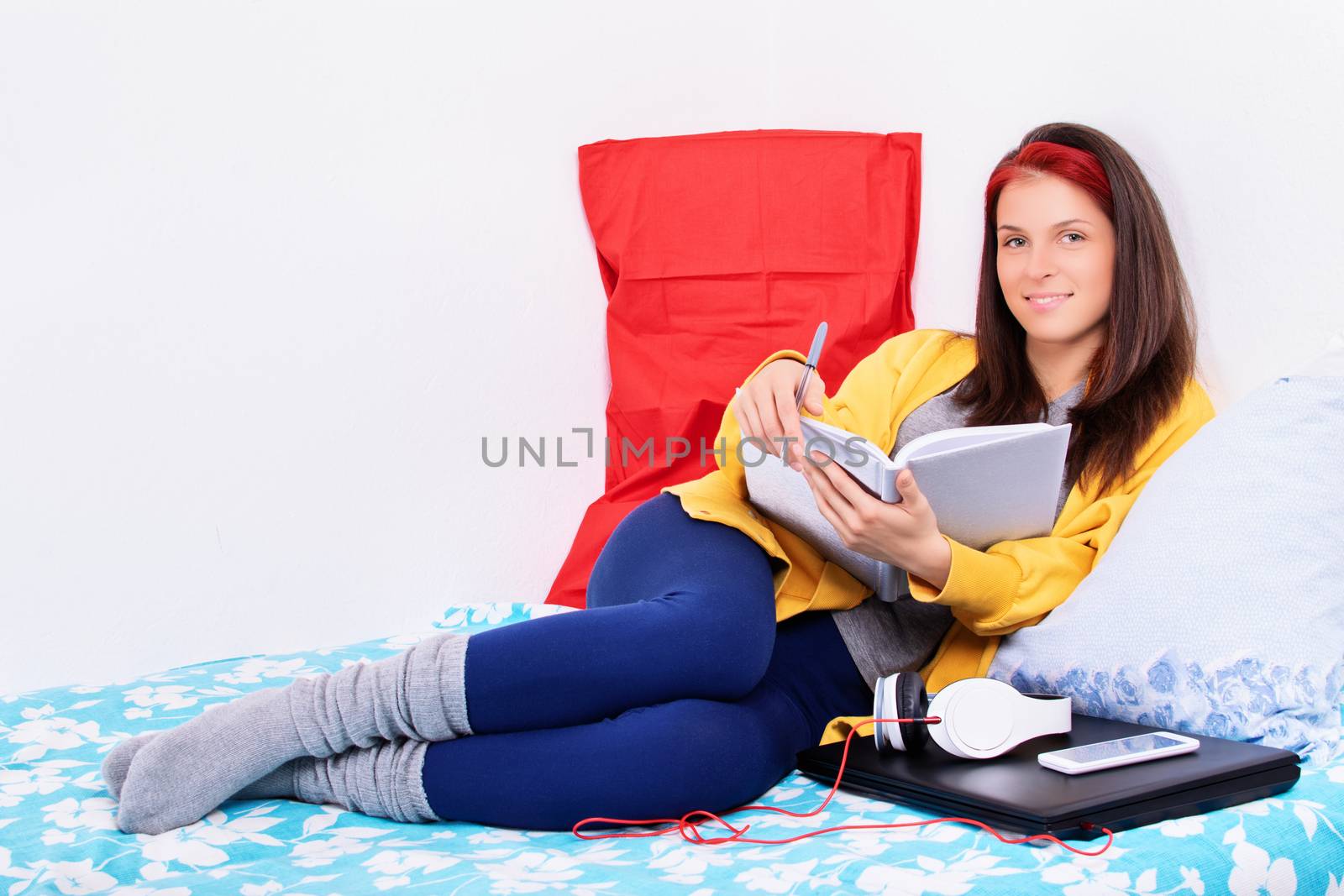 Beautiful young woman sitting comfortably on a bed in her bedroom holding a notebook, smiling and looking at the camera. Smiling girl relaxing in bed and making schedule for tomorrow.