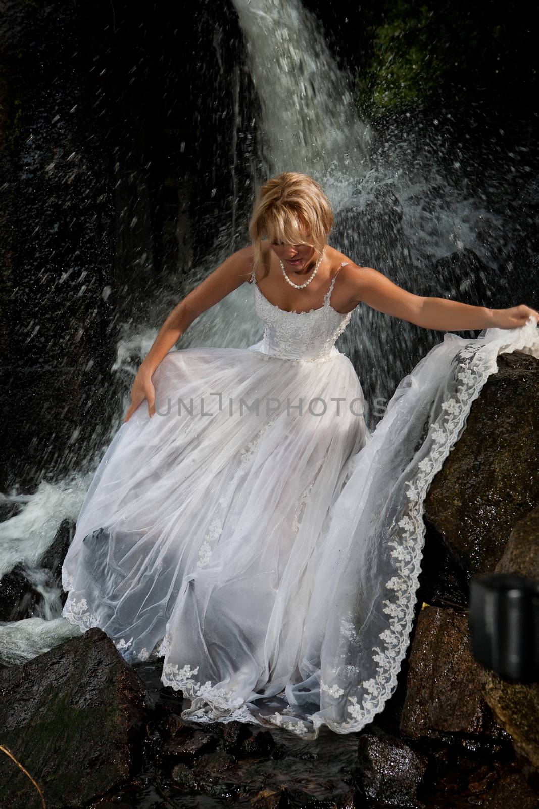 Young blonde woman in a white wedding dress near the waterfall