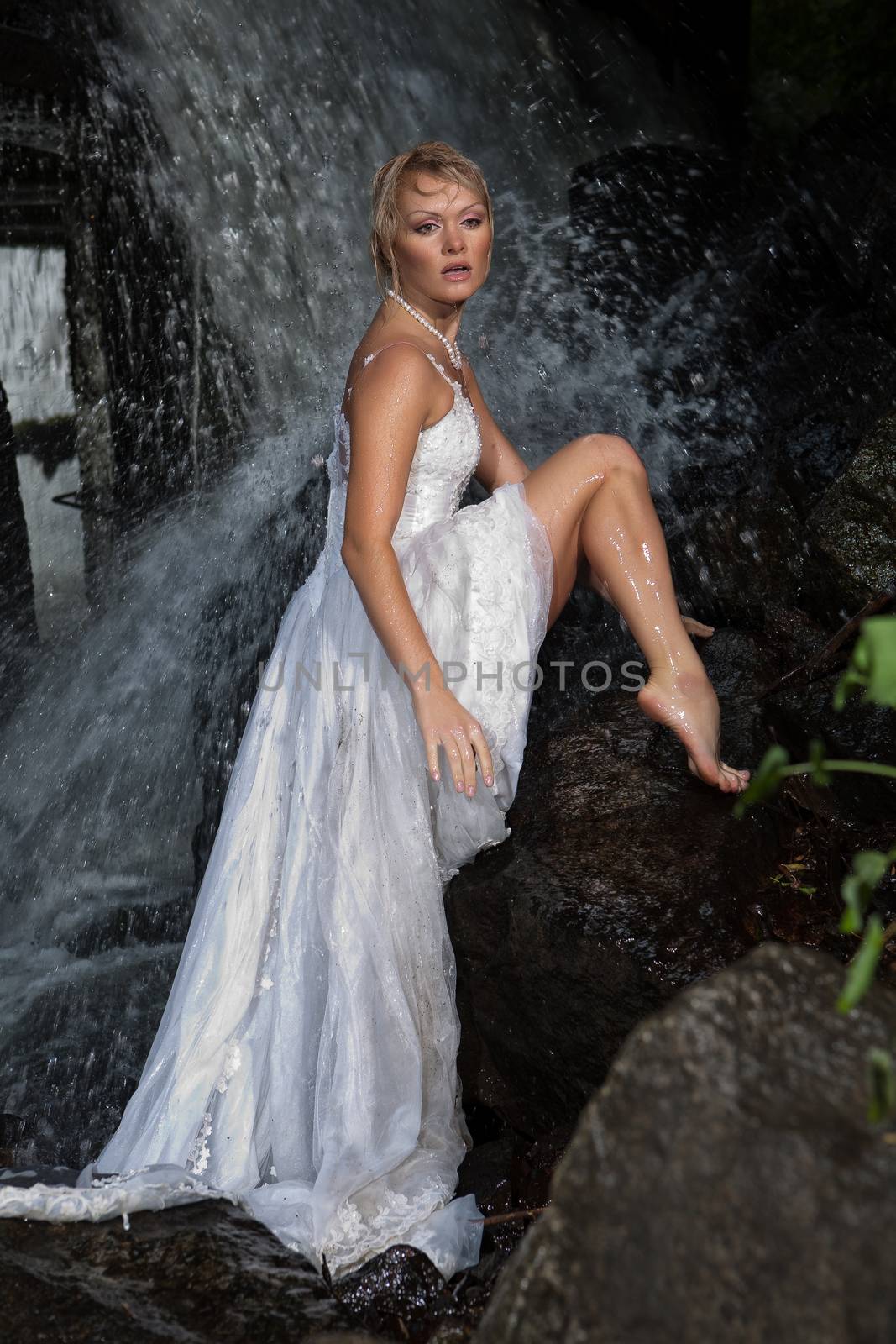 Young blonde woman in a white wedding dress near the waterfall