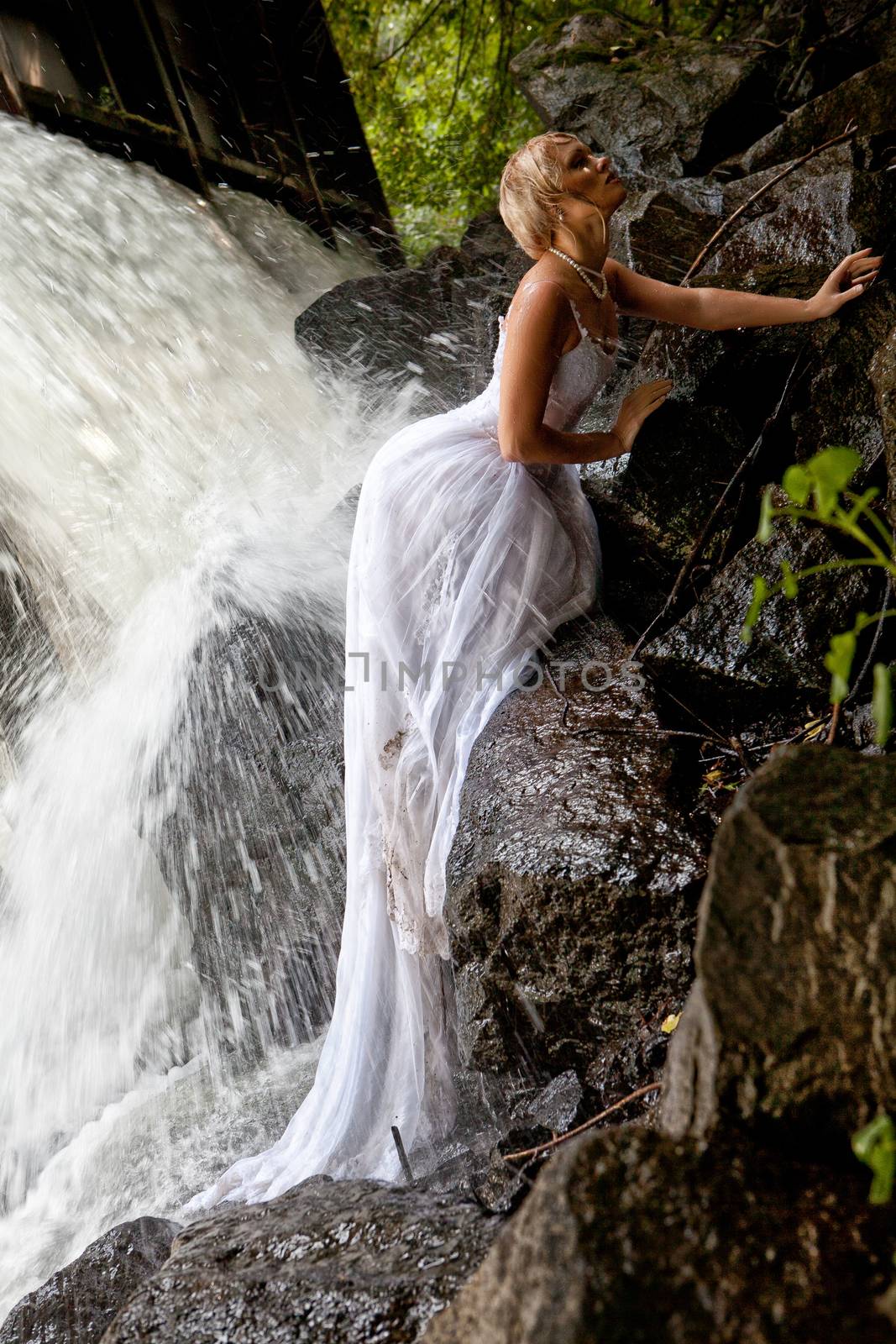 Young blonde woman in a white wedding dress near the waterfall