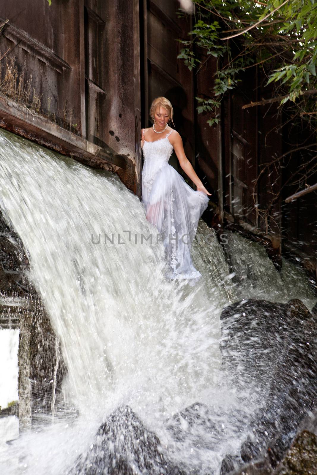 Young blonde woman in a white wedding dress near the waterfall