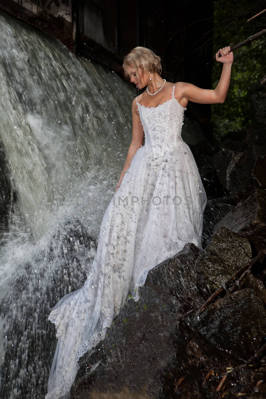 Young blonde woman in a white wedding dress near the waterfall