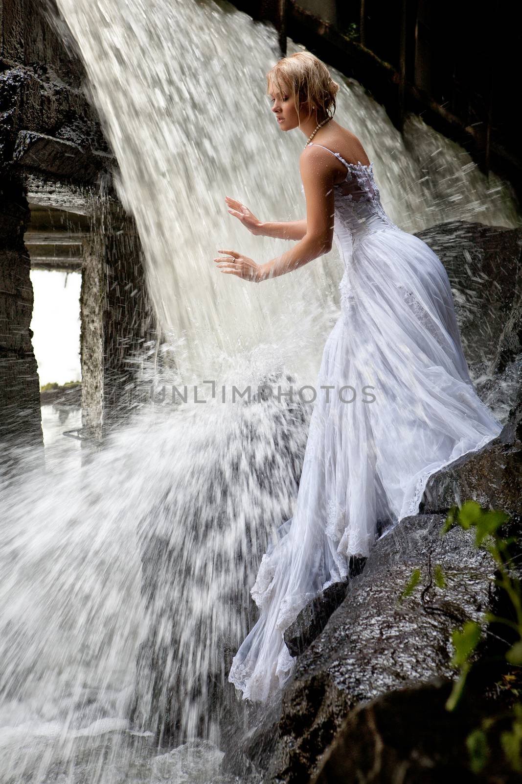 Young blonde woman in a white wedding dress near the waterfall