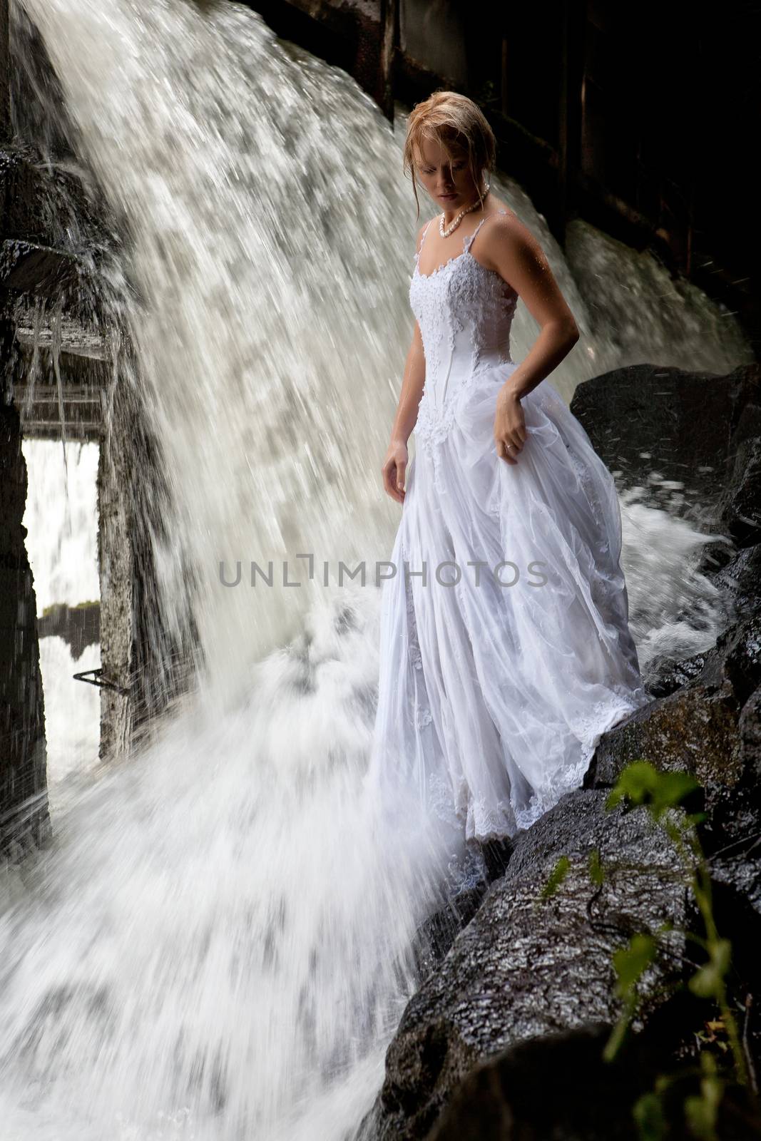 Young blonde woman in a white wedding dress near the waterfall