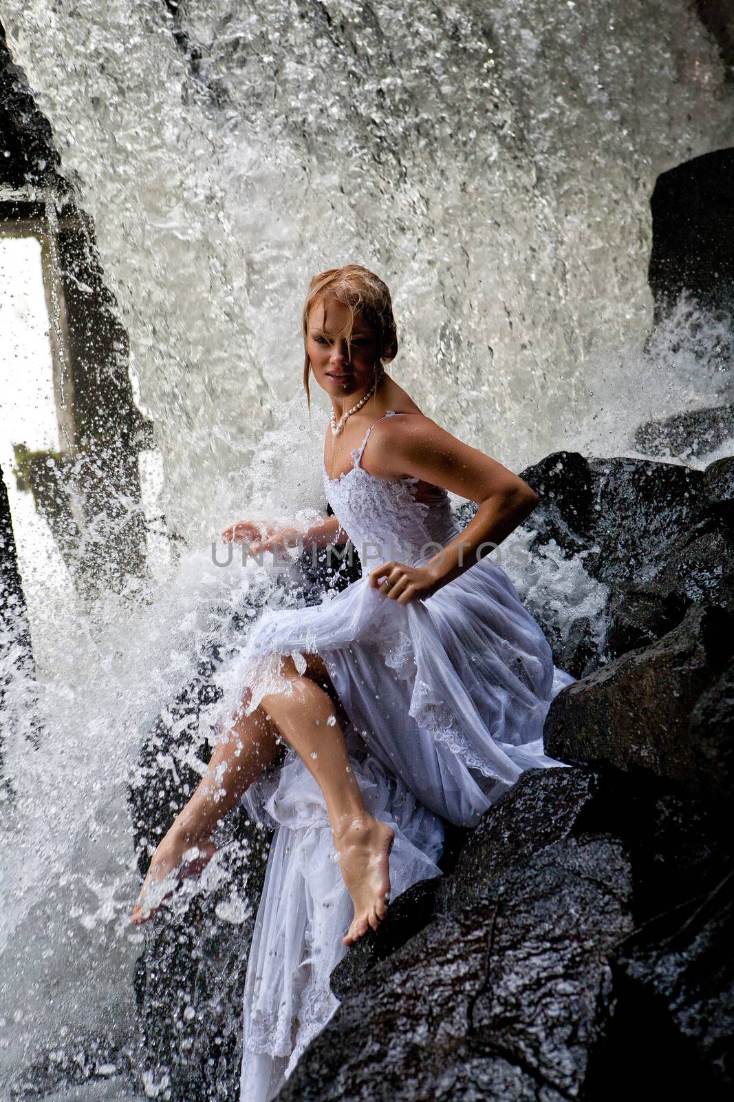 Young blonde woman in a white wedding dress near the waterfall