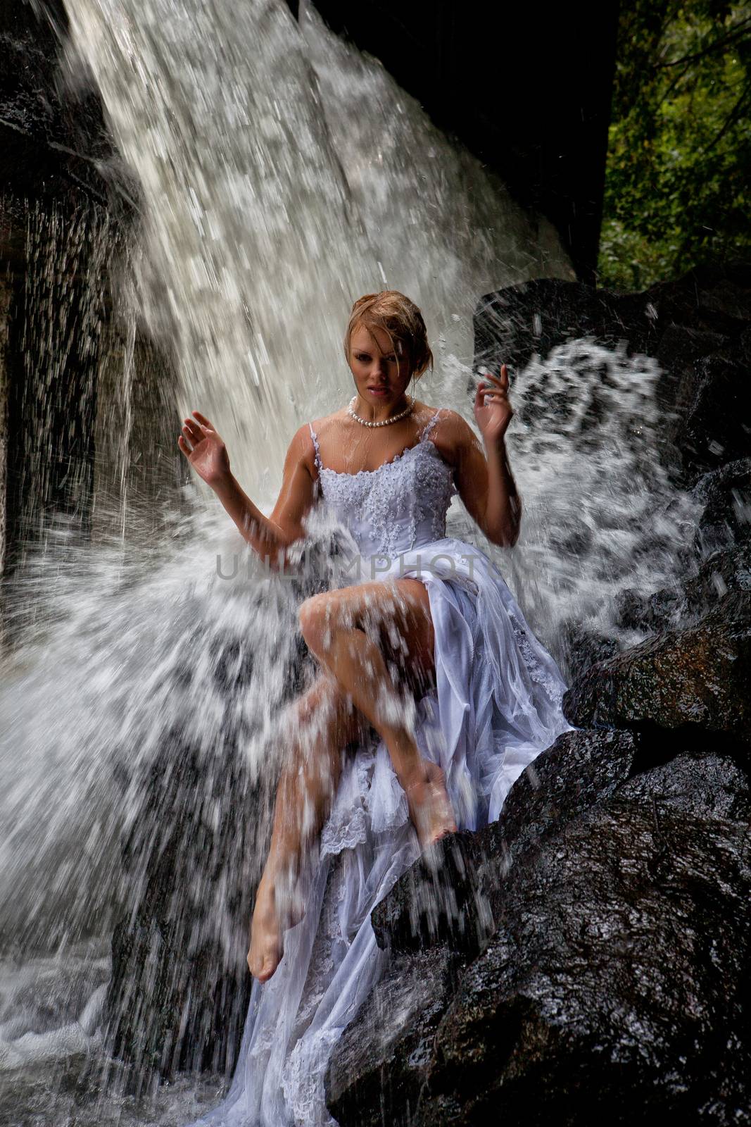 Young blonde woman in a white wedding dress near the waterfall