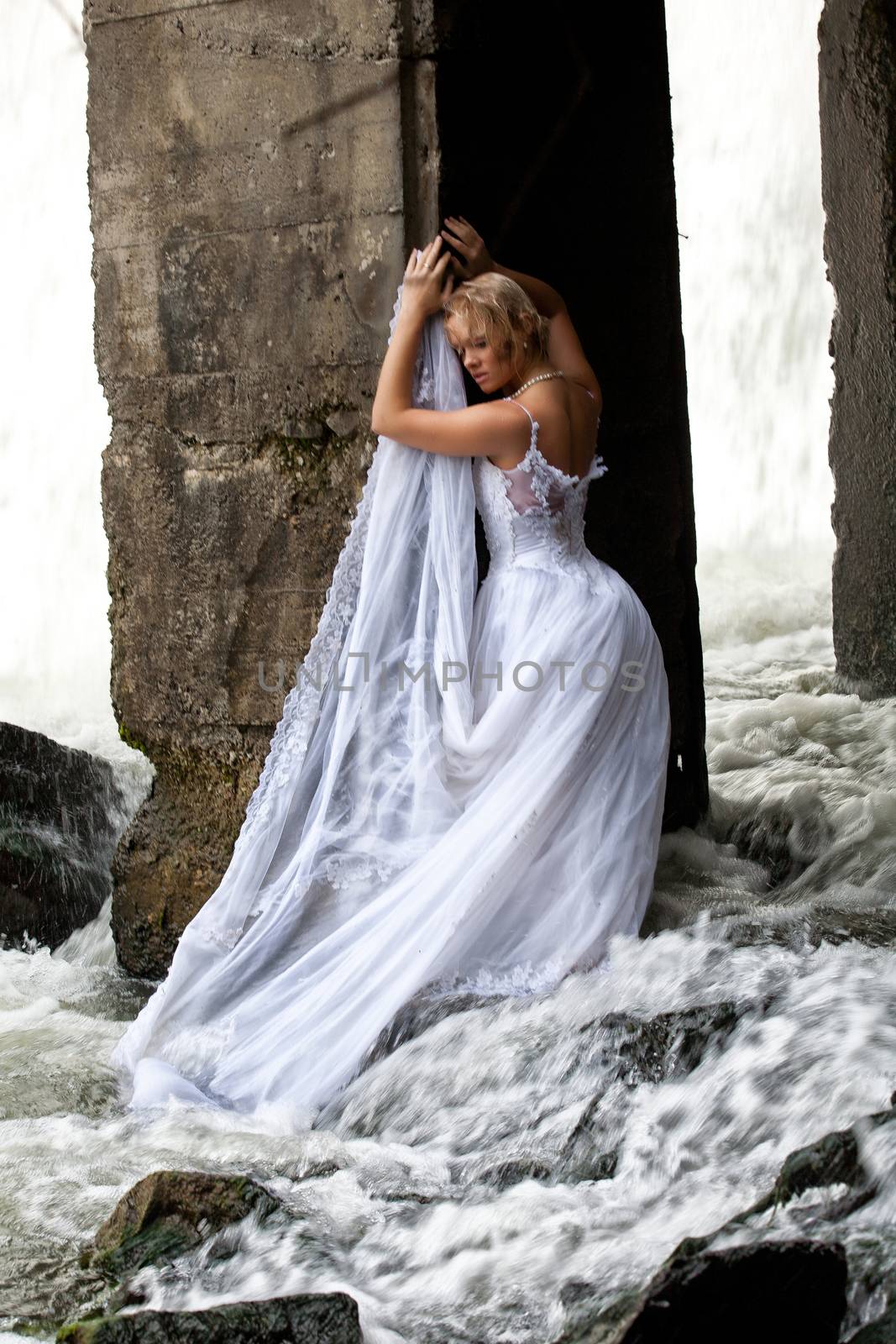 Young blonde woman in a white wedding dress near the waterfall