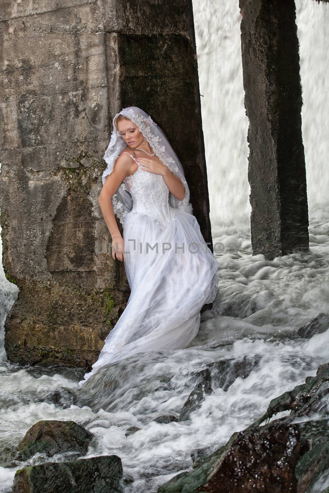 Young blonde woman in a white wedding dress near the waterfall