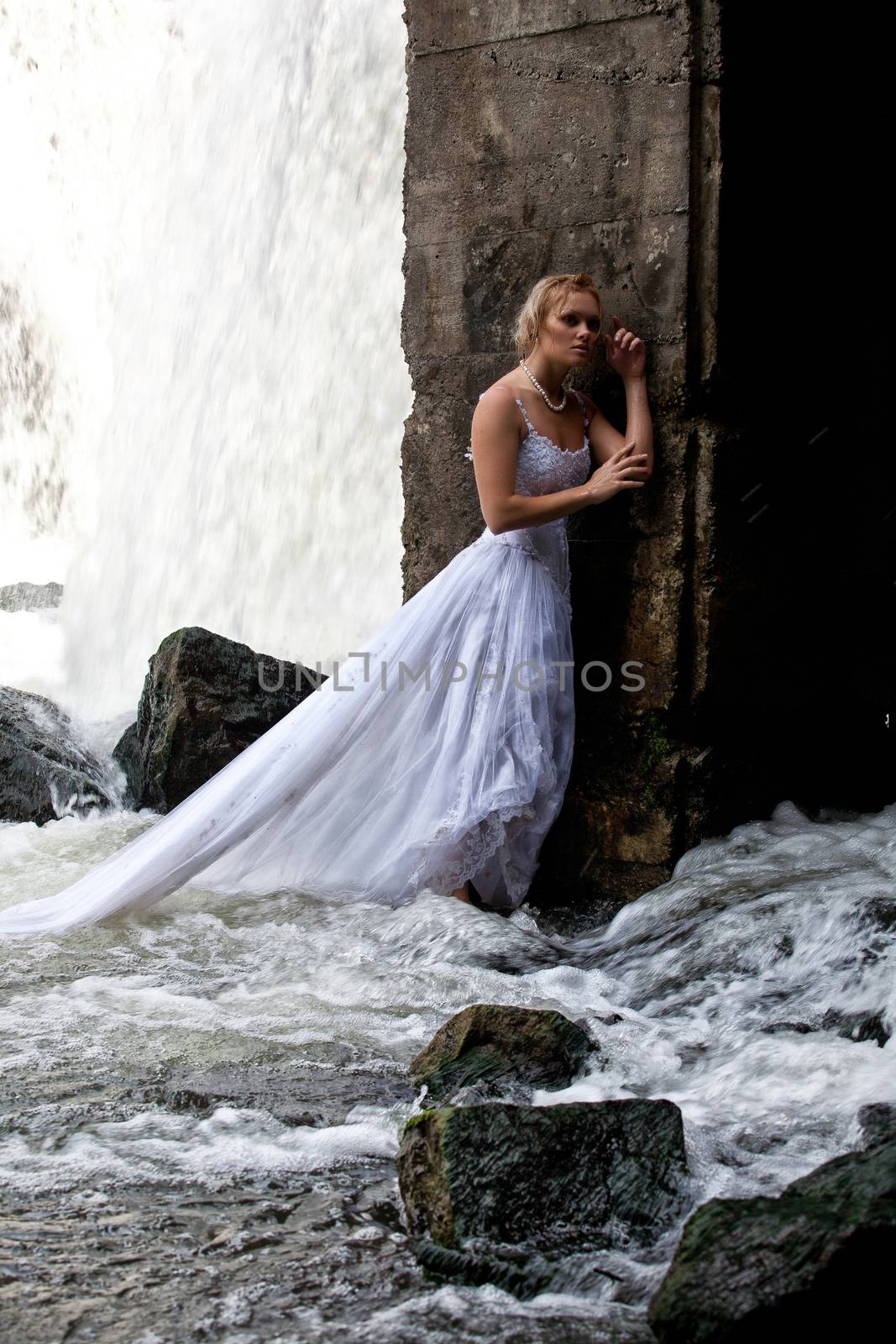 Young blonde woman in a white wedding dress near the waterfall