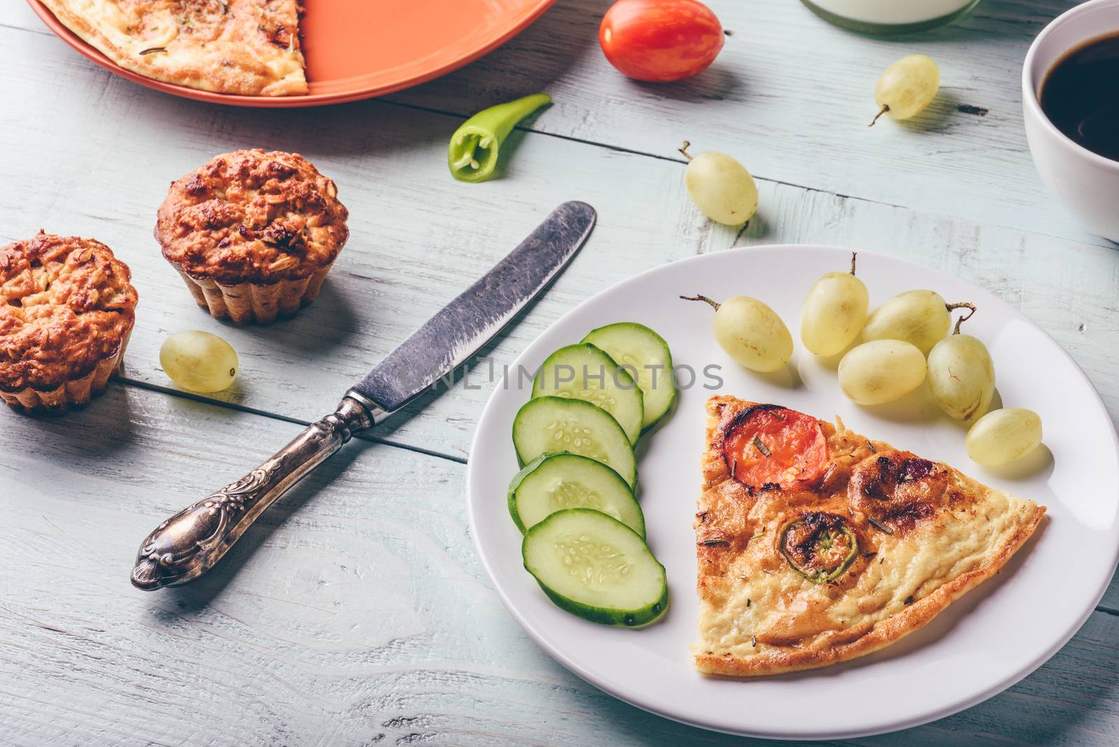 Slice of frittata with cup of coffee, grapes and muffins over light wooden background. Healthy eating concept.