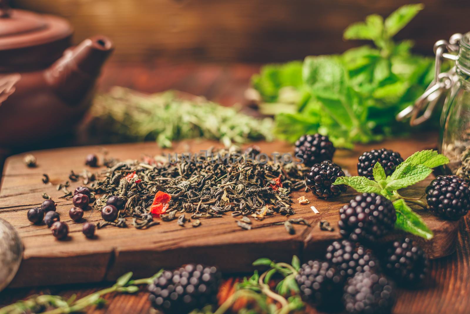 Heap of Dry Green Tea and Fresh Blackberries on Wooden Cutting Board. Bundles of Mint and Thyme Leaves. Clay Tea Pot.